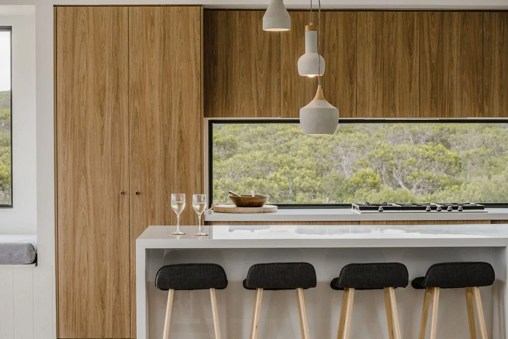 Modern kitchen with wood cabinets, a large window showing greenery outside, white countertops, a kitchen island with three black and wooden stools, two glasses of white wine, a wooden bowl, and hanging white pendant lights.