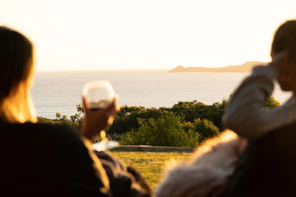 Two people sitting outdoors at sunset, with one holding a glass of wine, overlooking the ocean and a distant landmass.