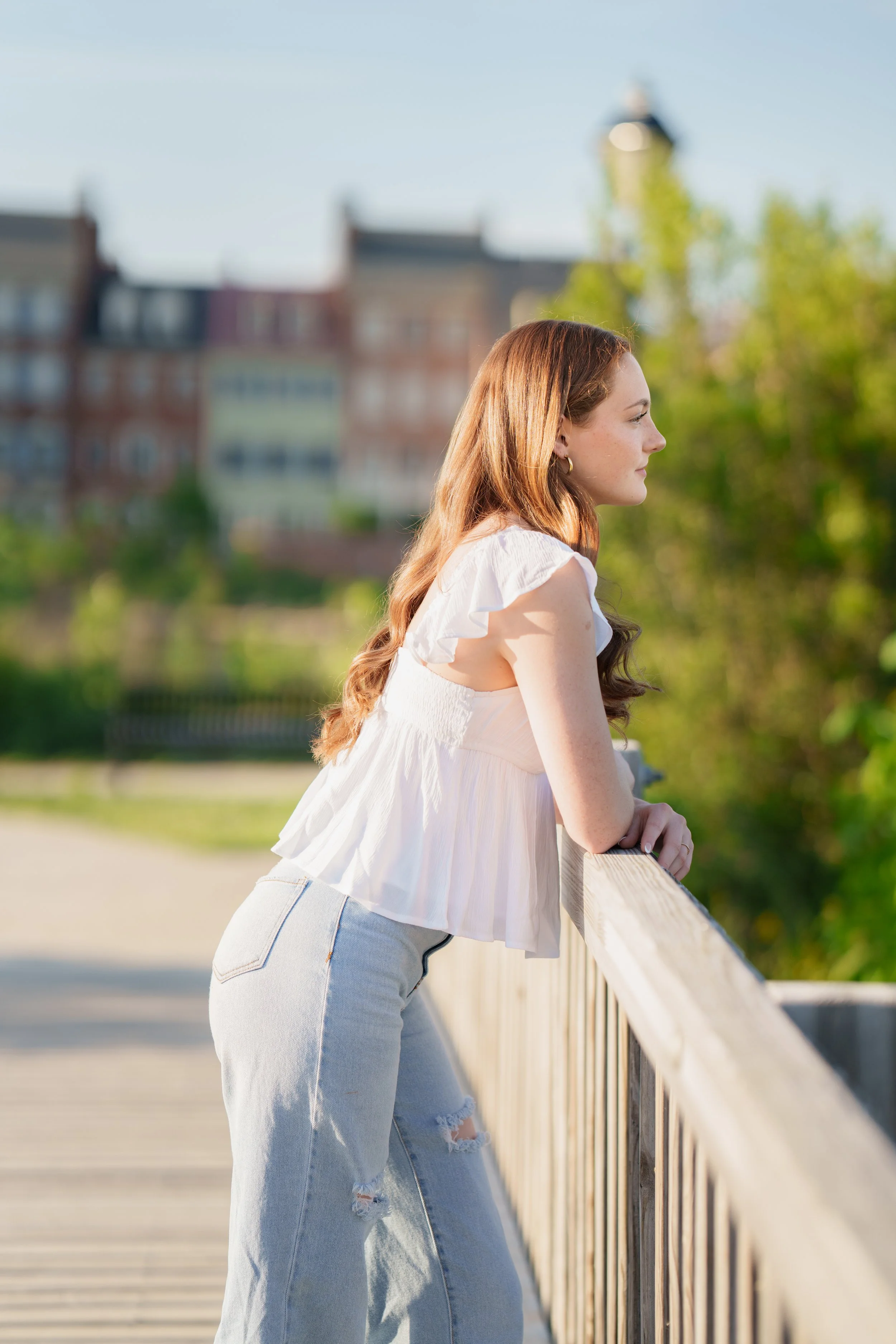 Young woman leaning on a railing outdoors, wearing a white top and ripped jeans, with a blurred background of trees and buildings.