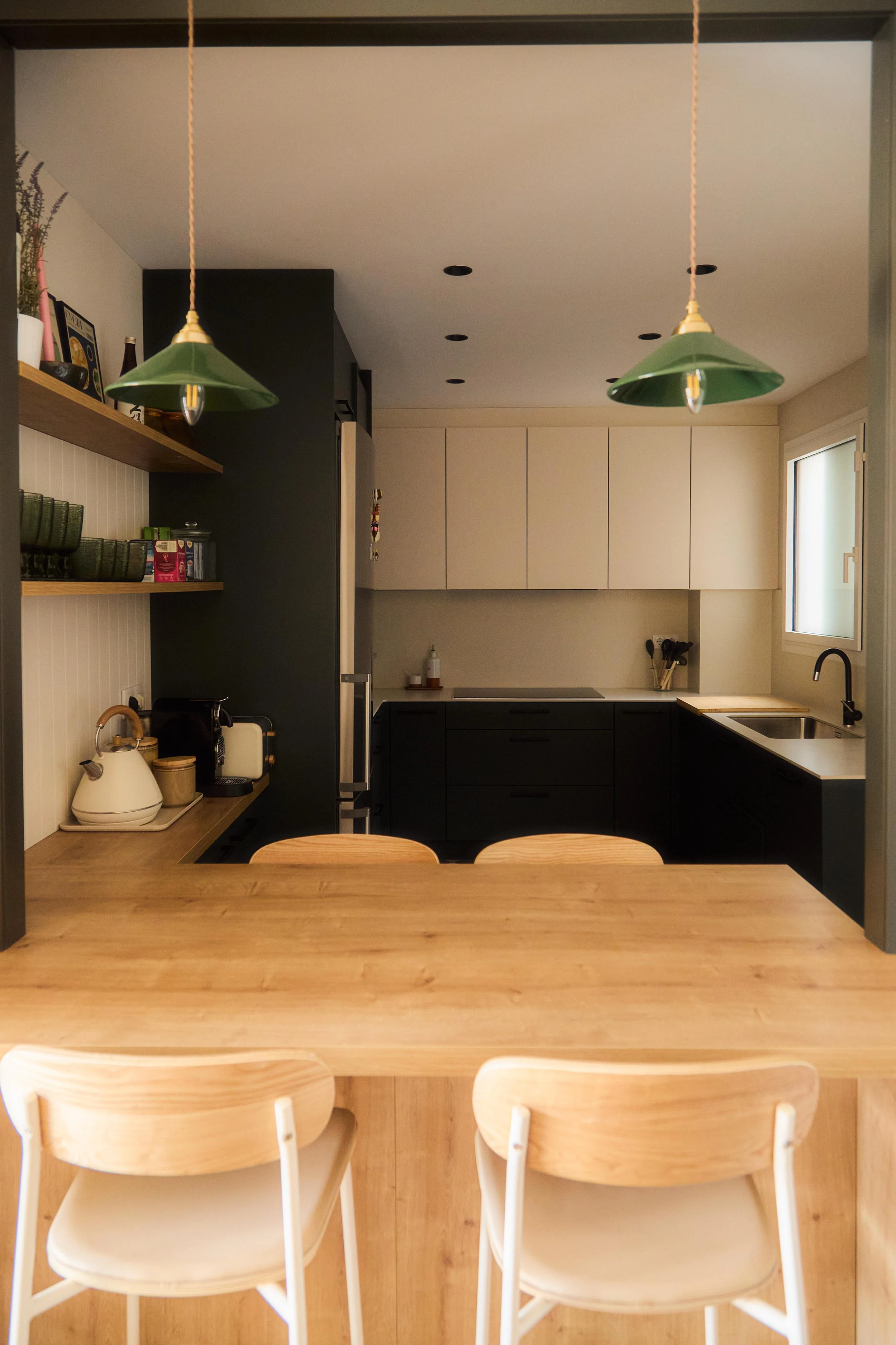 Cocina moderna con mesa de madera, sillas blancas, gabinetes blancos y negros, estantes con platos y utensilios, lámparas verdes, ventana por la que entra luz natural.