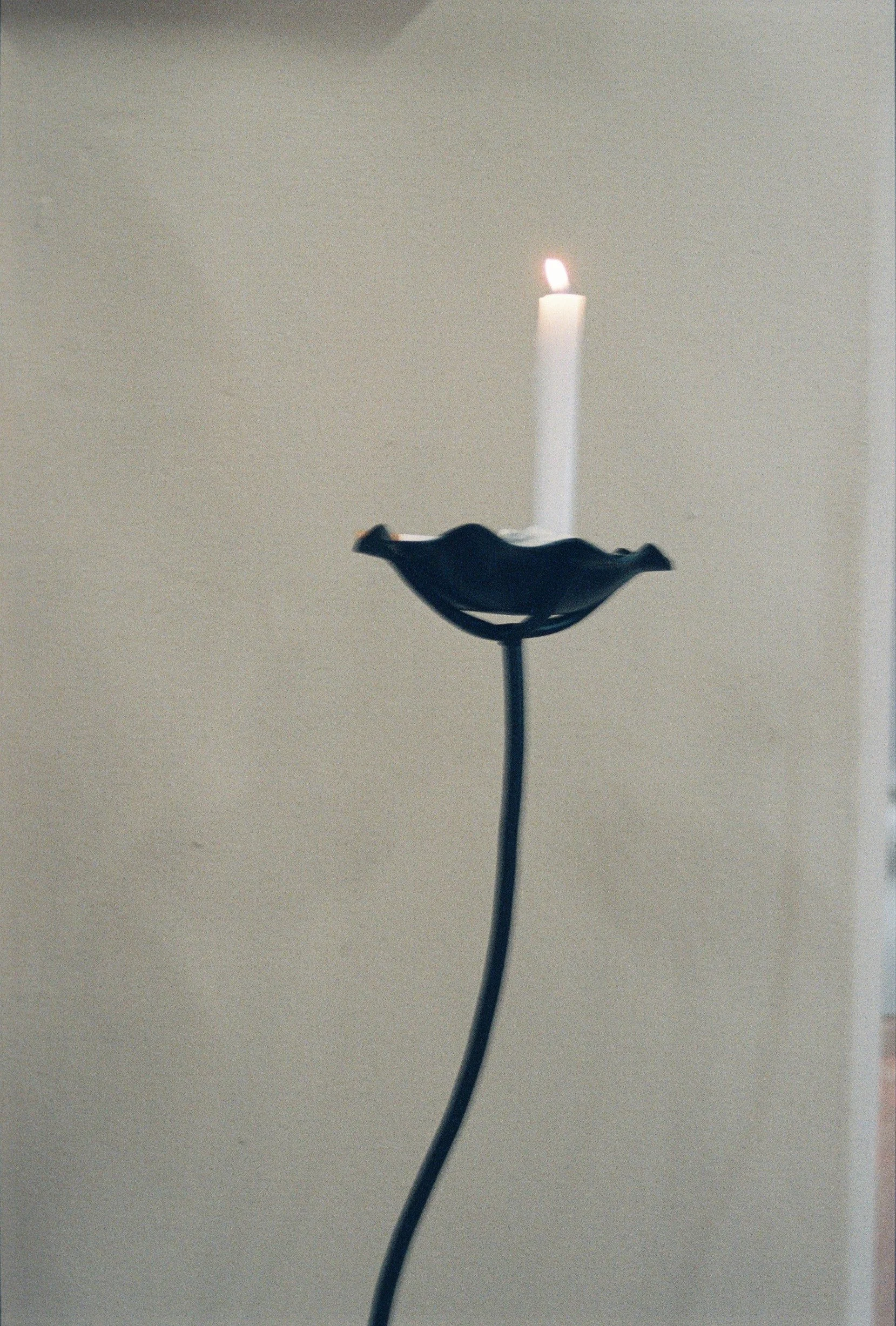 Vela encendida en un soporte artístico en forma de flor. La vela está en una base negra y la imagen tiene un fondo simple y claro.
