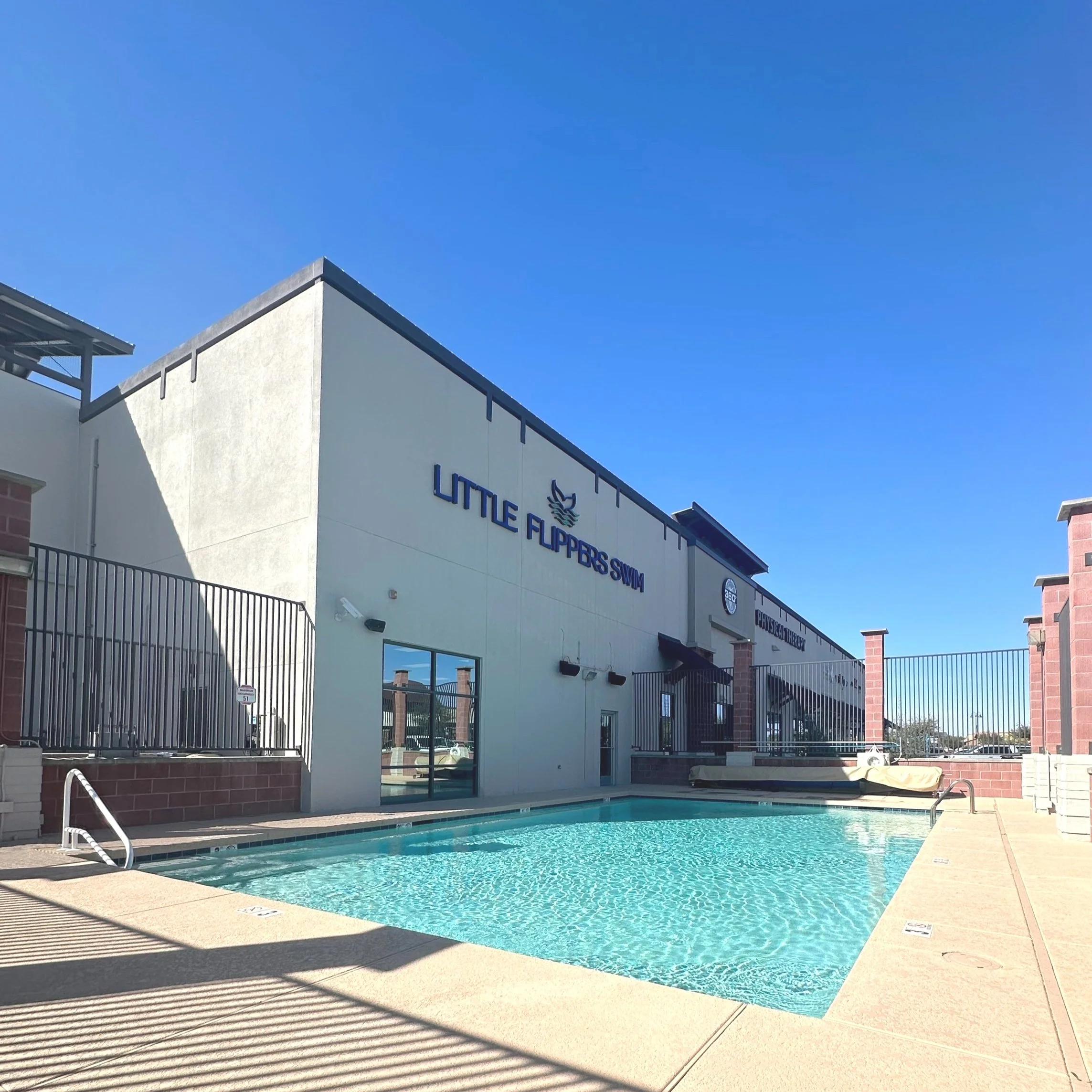 Outdoor pool area at Little Flippers Swim's Queen Creek Marketplace location with clear water and adjacent building under a blue sky.