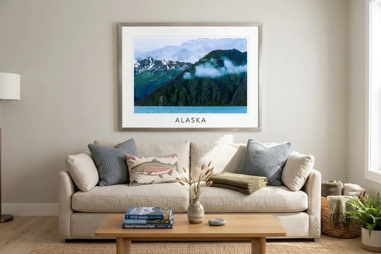 Living room with a beige sofa, gray pillows, and a framed landscape photograph of mountains labeled 'Alaska' hanging on the wall, a wooden coffee table with books, a vase with dried plants, and a window on the right.