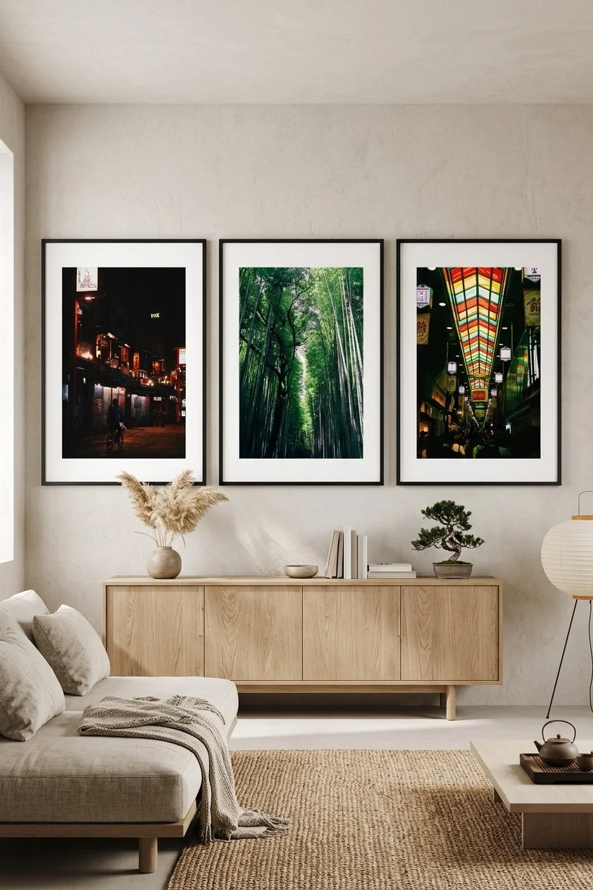 Living room with three framed photographs of city scenes at night, a forest, and indoor shopping mall hang above a wooden sideboard. Decor includes a beige sofa with pillows and a blanket, a vase with pampas grass, a bonsai tree, books, and a floor lamp with a paper shade.