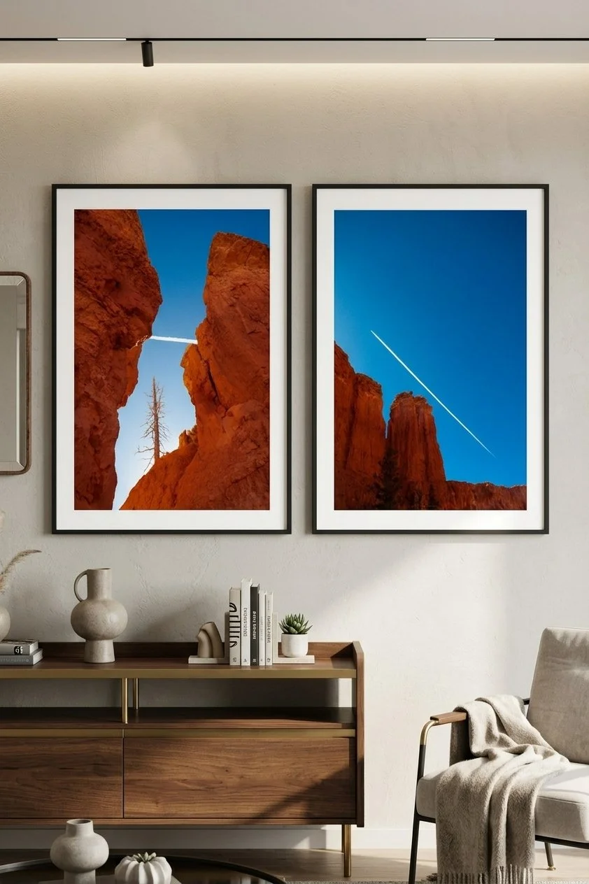 Two framed landscape photographs of red rock formations in a desert, with one photo showing a rock canyon under a lighthouse streaked sky and the other featuring tall canyon walls with a contrail in the blue sky, hanging above a mid-century modern wooden sideboard in a cozy living room.