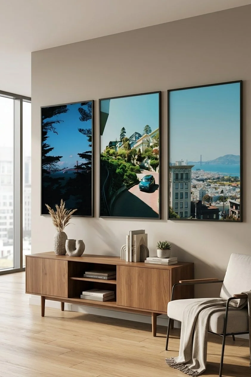 Interior of a modern living room with a wooden sideboard decorated with vases, books, and a small plant, a white chair with a throw blanket, and three large framed pictures of city views and landscapes hanging on the wall.