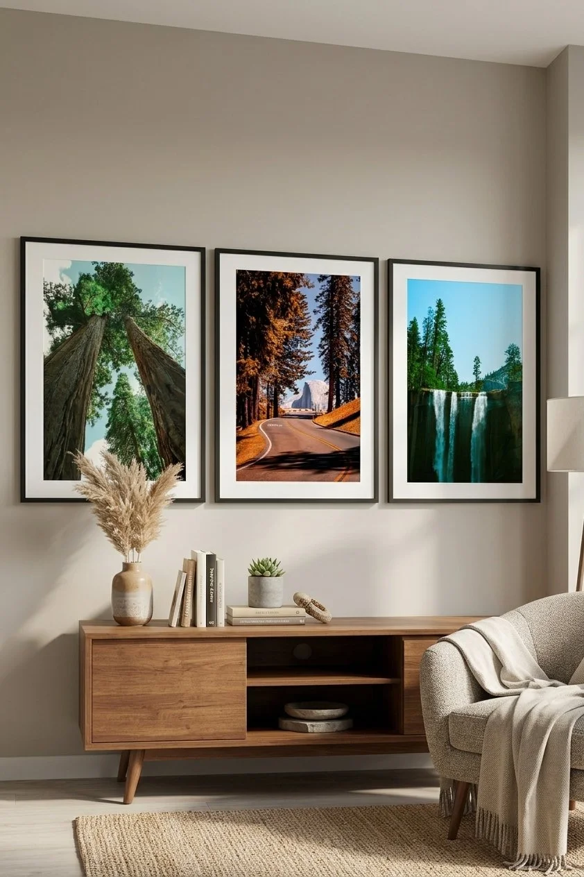 Three framed landscape photos of trees, a road through a forest, and a waterfall hang above a wooden console table with decorative items, including a vase with dried pampas grass, books, and a small potted succulent, in a neutral-colored living room.