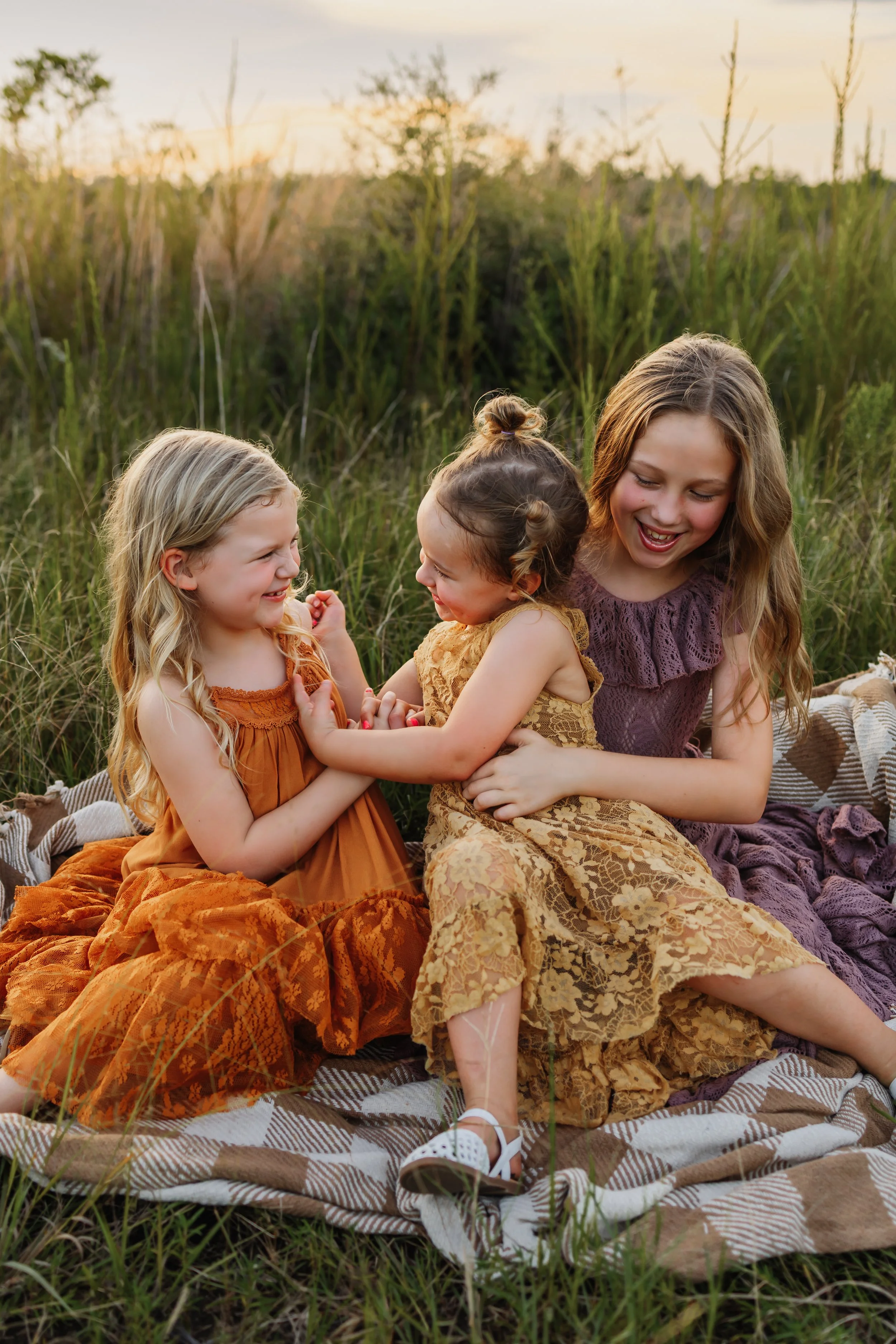 Three young sisters tickling each other while sitting on a blanket in a field near Pensacola, FL during a photoshoot with Kristen Coberly Photography