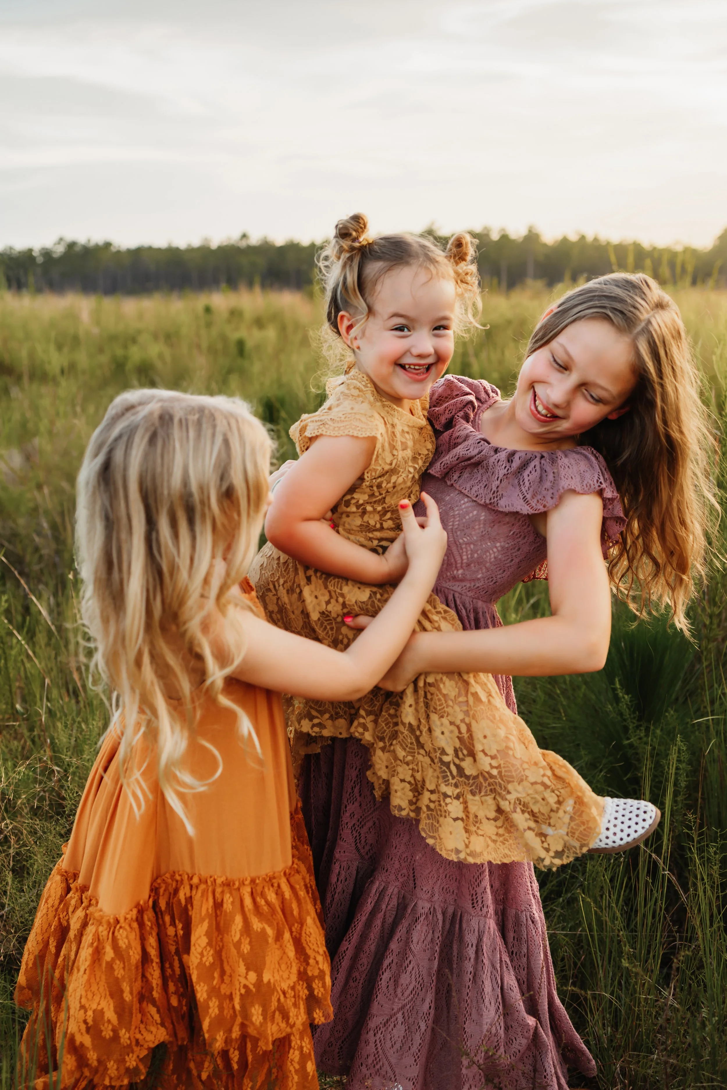 Two sisters laughing while the middle sister tickles them in a field near Pensacola, FL during a session with Kristen Coberly Photography