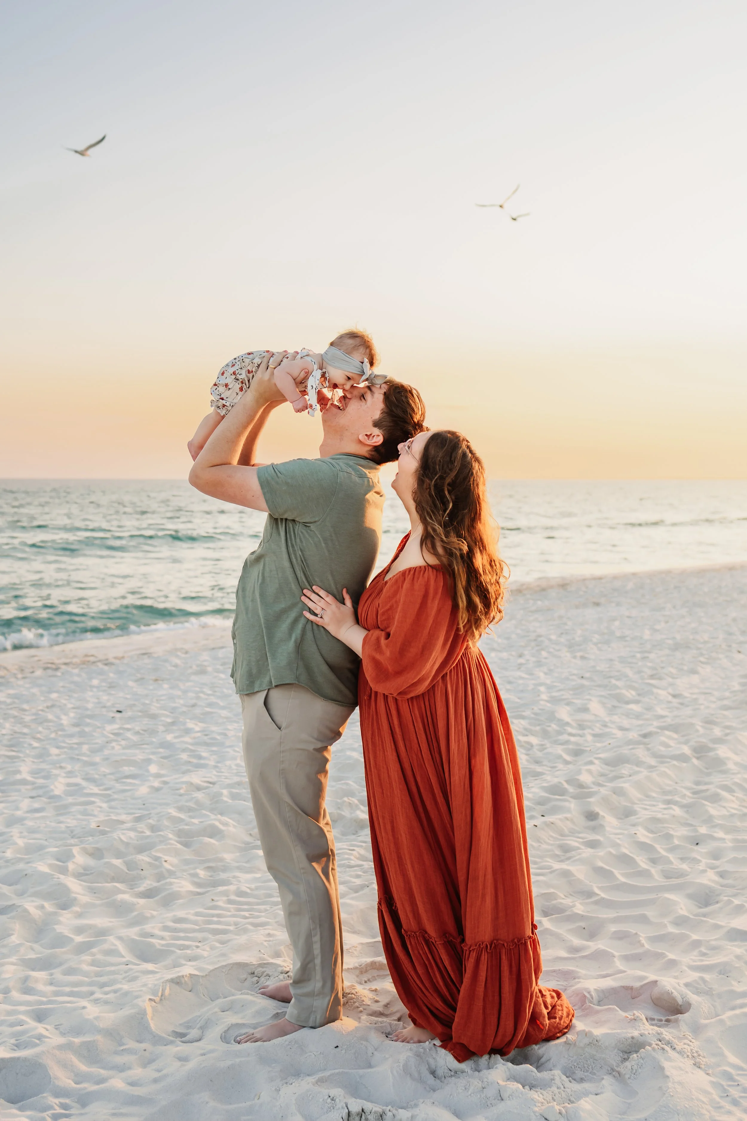 Mom hugging her husband from behind as her husband brings their 9 month old to his nose with the Gulf of America behind them and the white sandy beach of Pensacola under their toes during a session with a Pensacola Beach Photographer