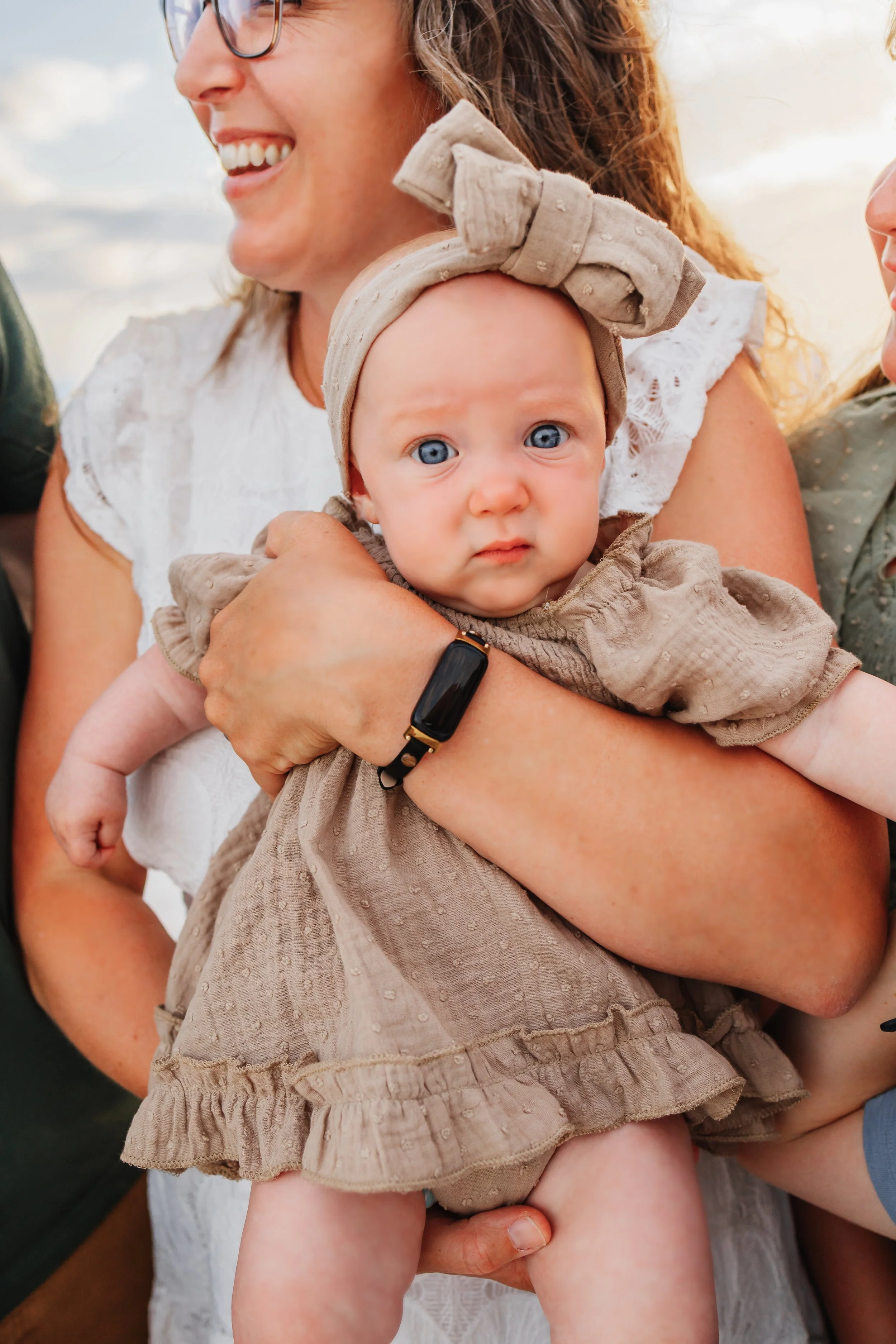 Baby girl in tan dress looking at the camera while in her mother's arms during a photo session on Pensacola Beach with Kristen Coberly Photography