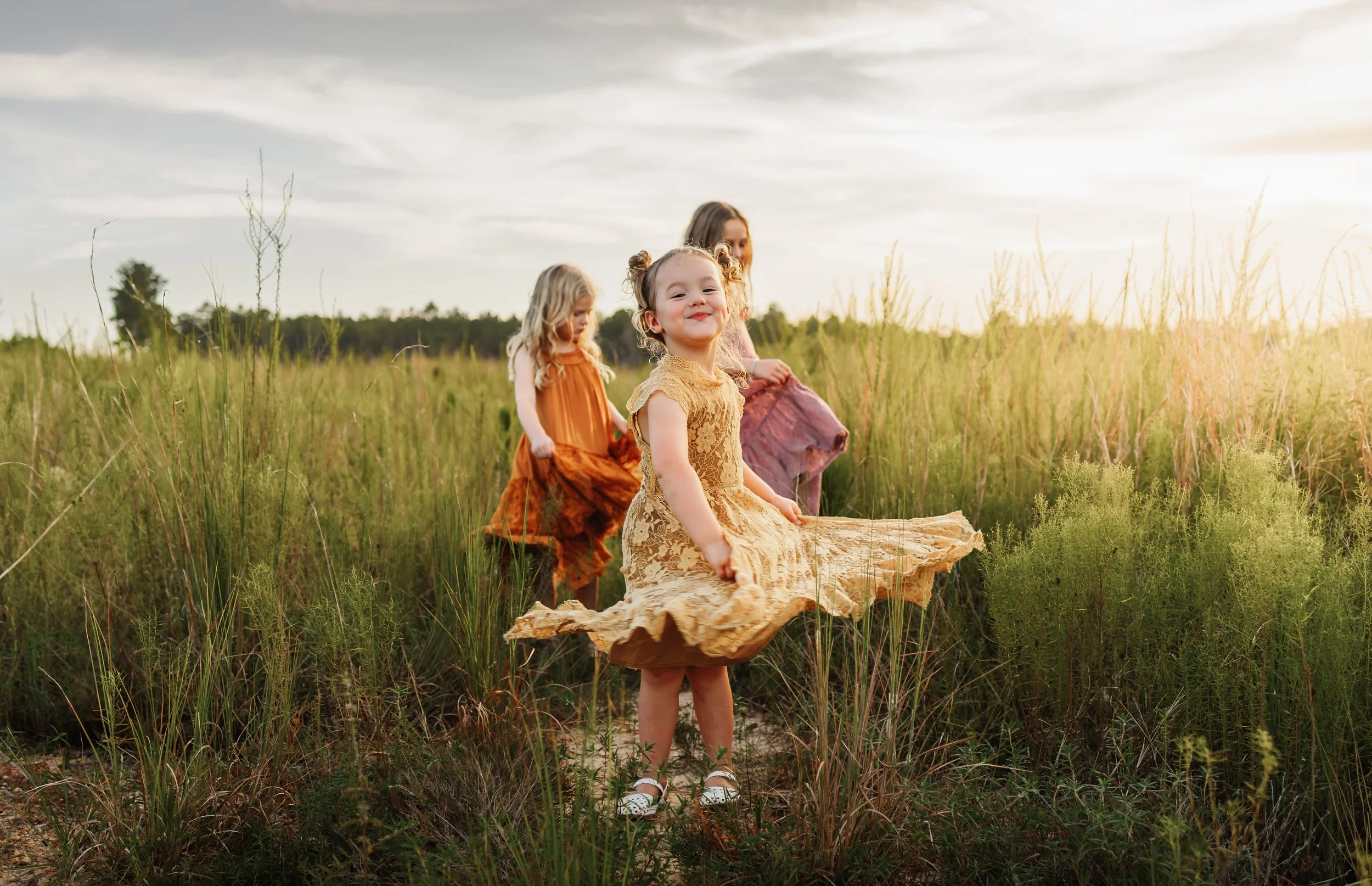 A little girl in a yellow dress twirls in a field near Pensacola, FL with her older sisters in the background during a photoshoot with the best photographer in Pensacola, Florida Kristen Coberly Photography