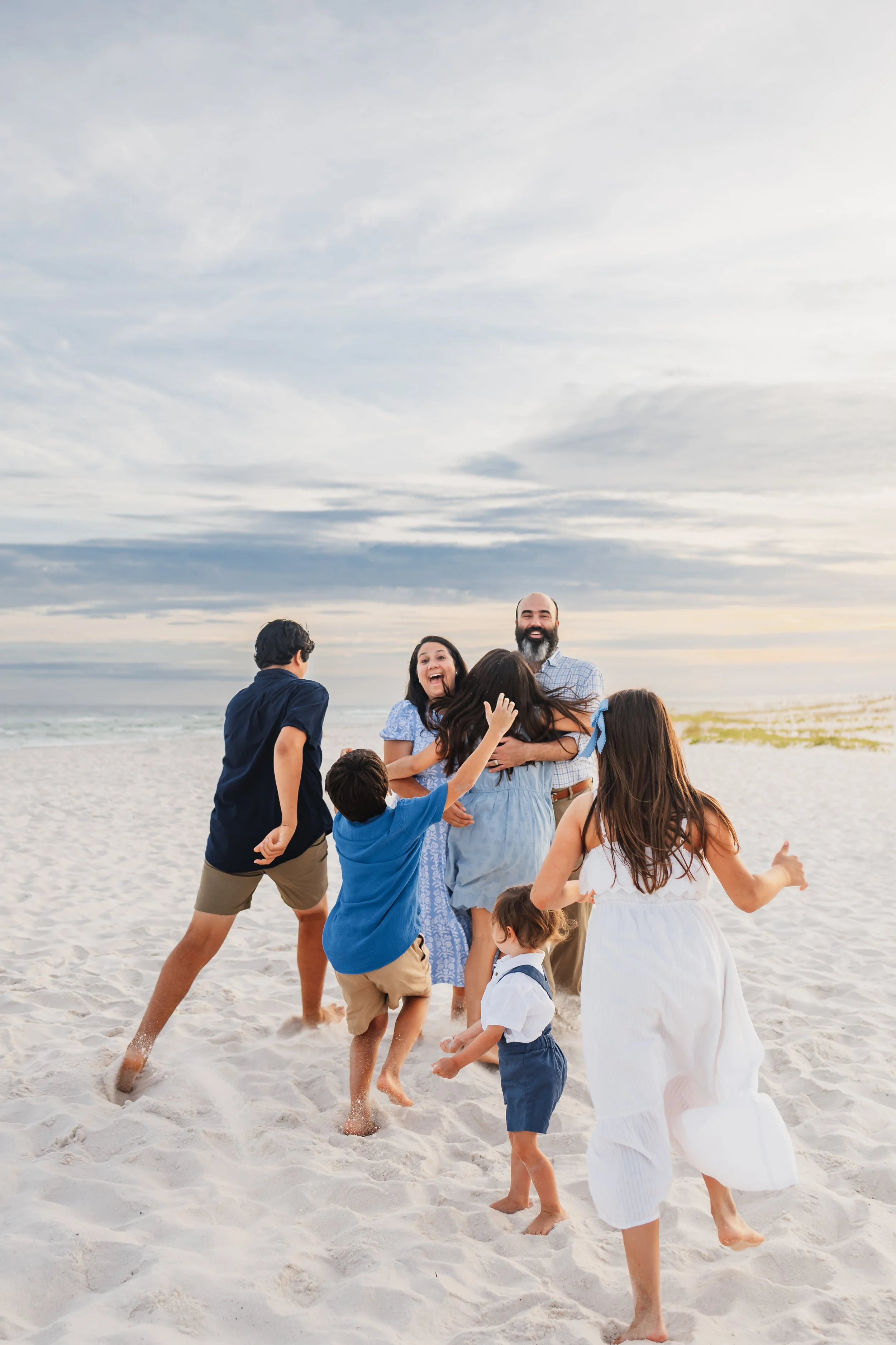 Six children wearing blue tones running toward there parents in a surprise attack hug on the white sands of Pensacola Beach during a photoshoot with Kristen Coberly Photography