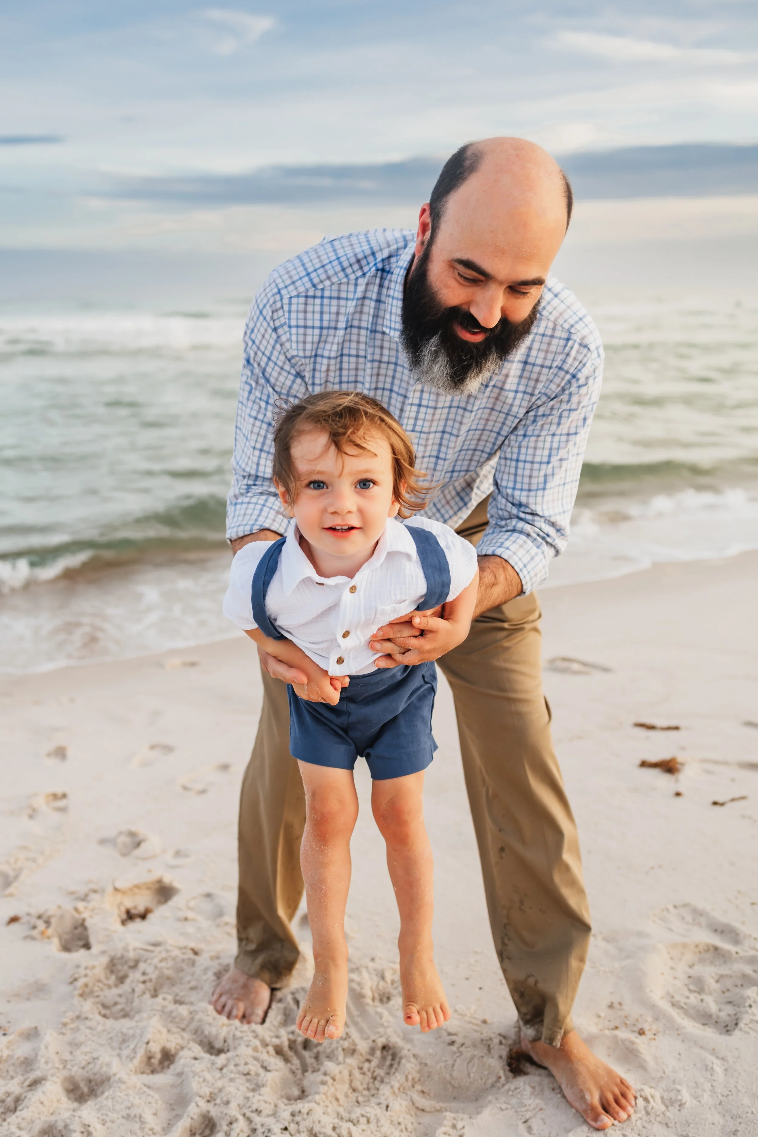 Dad lifting up his 2 year old son while playing with him by the water on Pensacola Beach during a photoshoot with Kristen Coberly Photography.