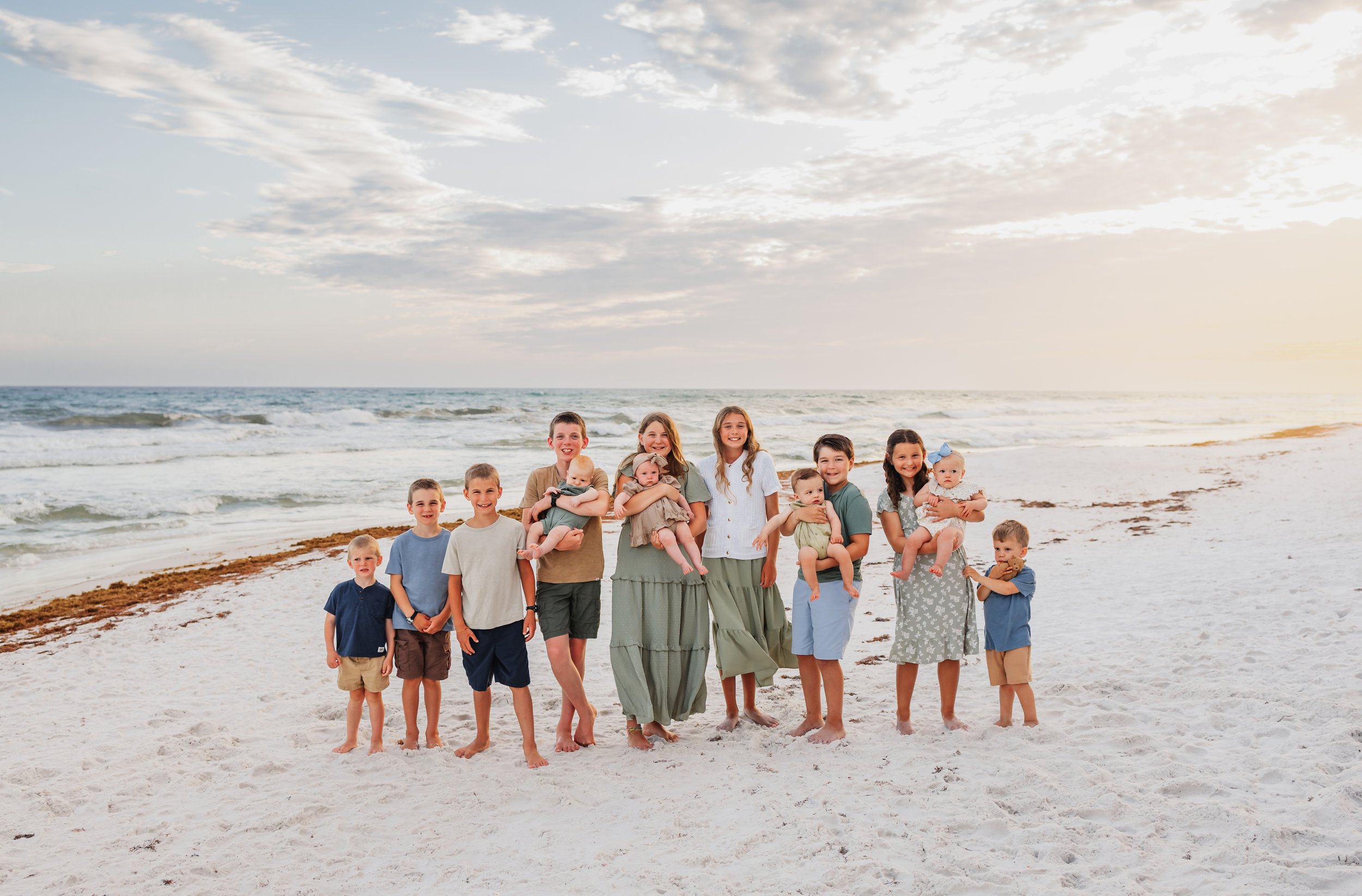 All thirteen grandkids lined up in a line smiling at the camera while standing on the white sand of Pensacola Beach during a session with Kristen Coberly Photography