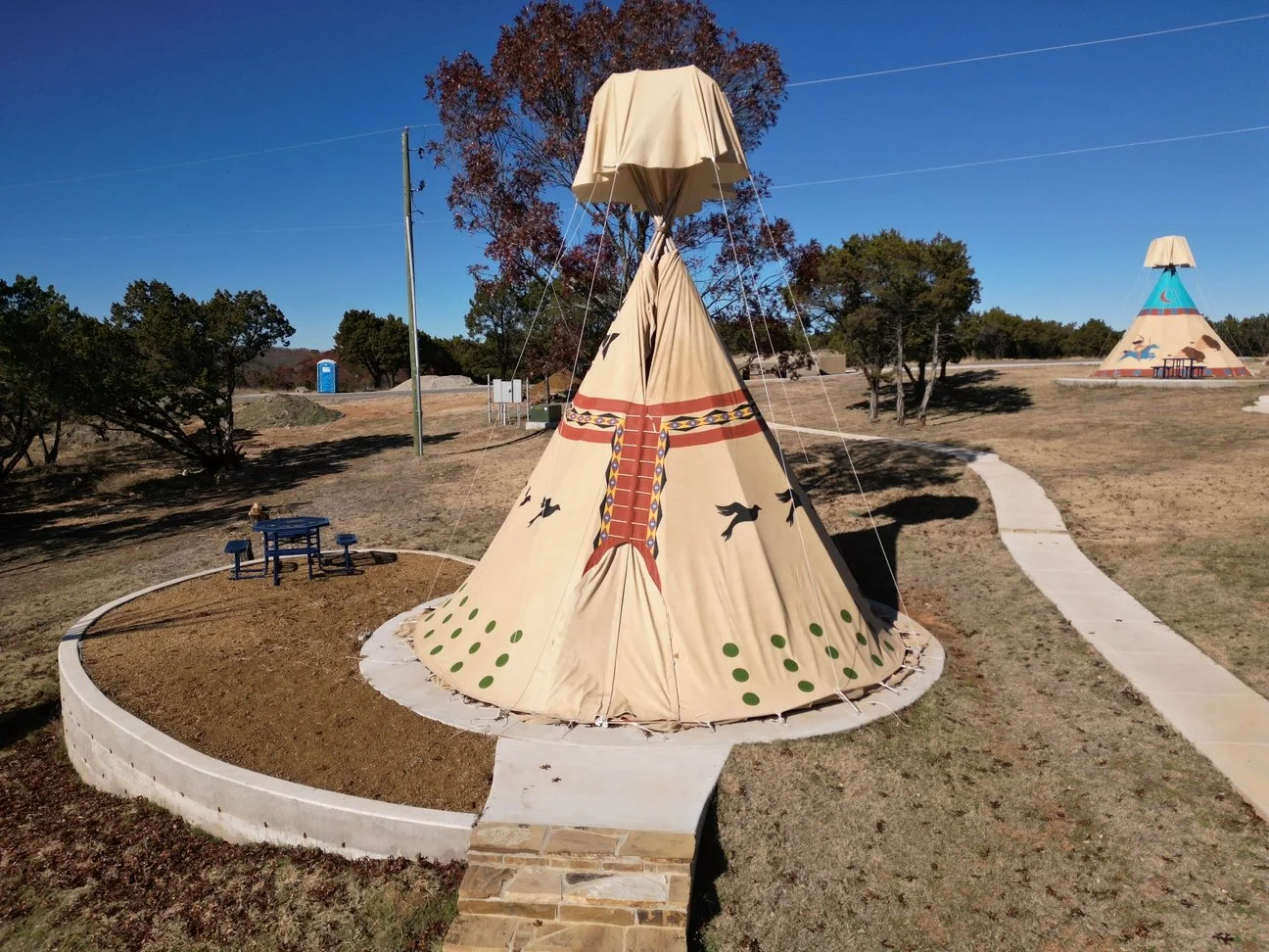 A teepee tent with a decorative design sits on a wooden platform outdoors during sunset, with two chairs, a small table, and a lantern in front.