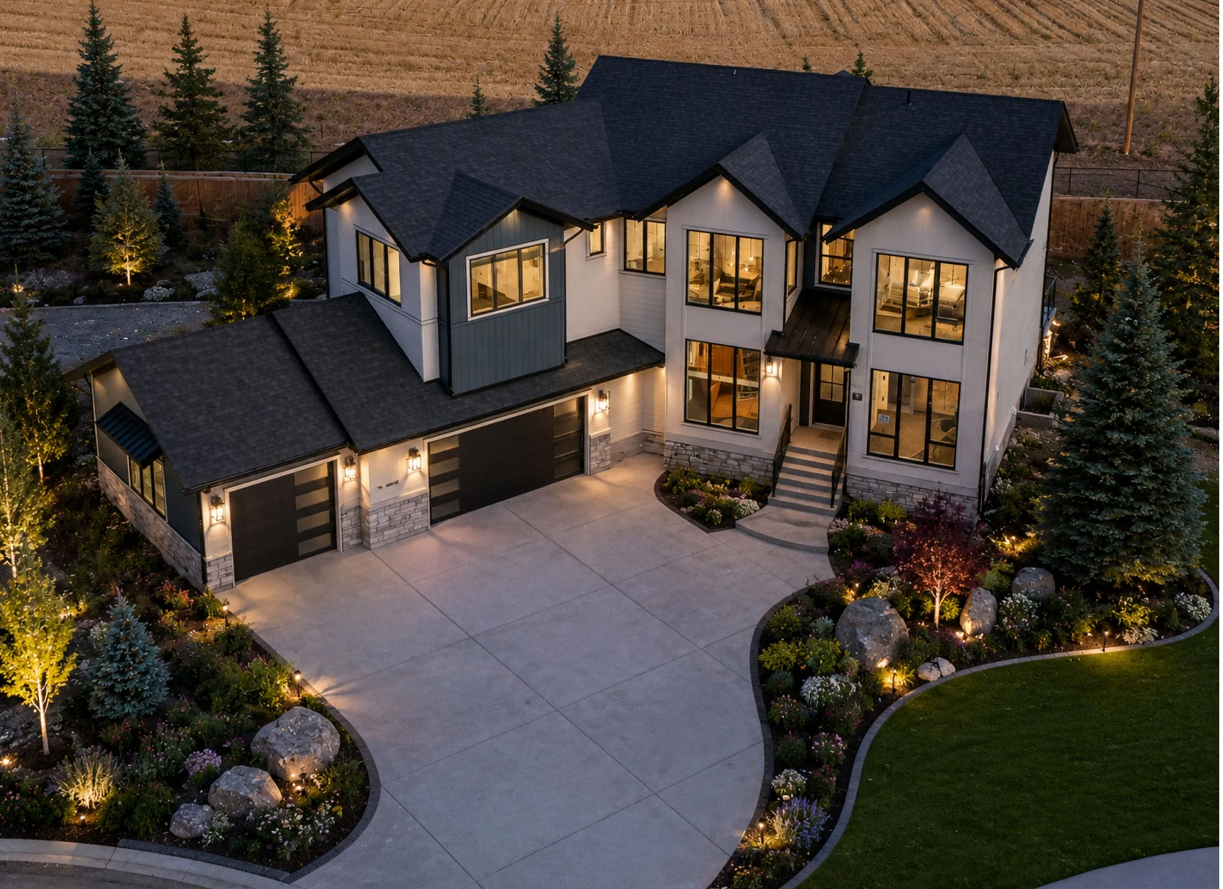 A large modern house with multiple stories and large windows illuminated from inside, surrounded by trees and landscaped garden at dusk.