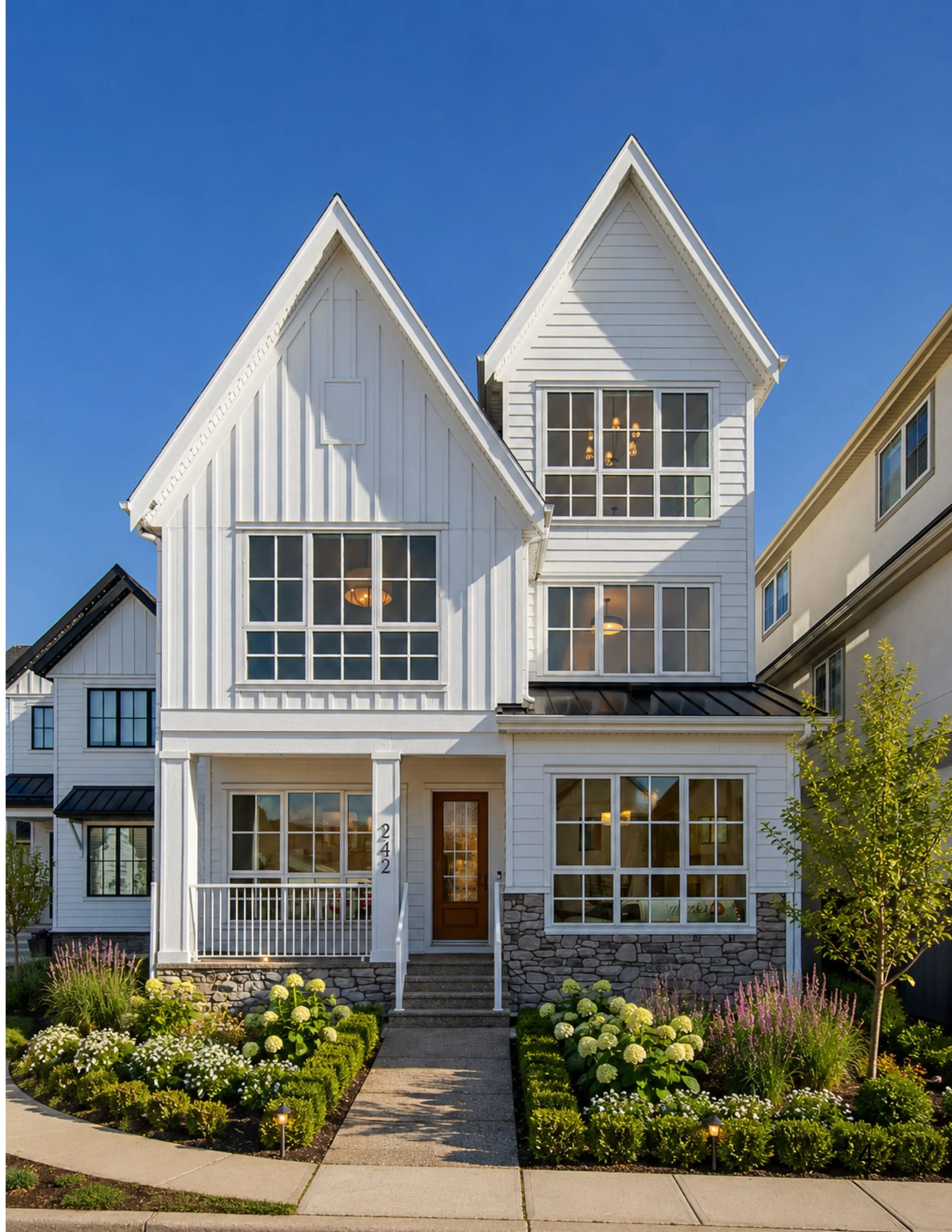 White multi-story house with large windows, a front porch, and a landscaped garden with various flowering plants in the front yard.