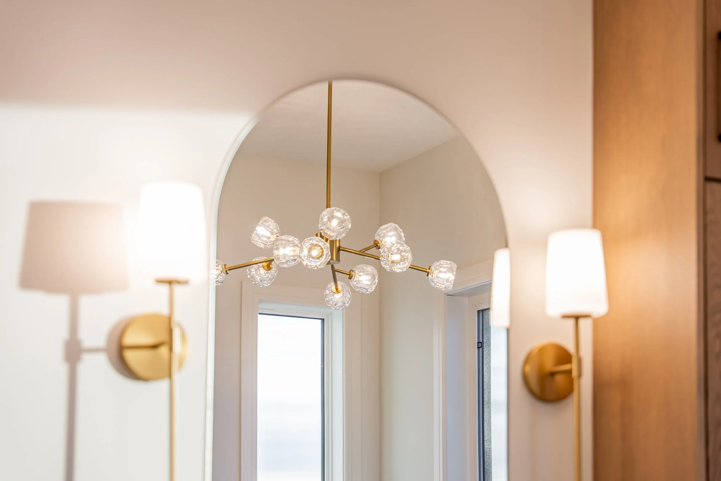 A room with a modern chandelier with glass globes hanging from a brass fixture. Two wall-mounted lamps with white shades are on either side, and there is a window with a white frame.