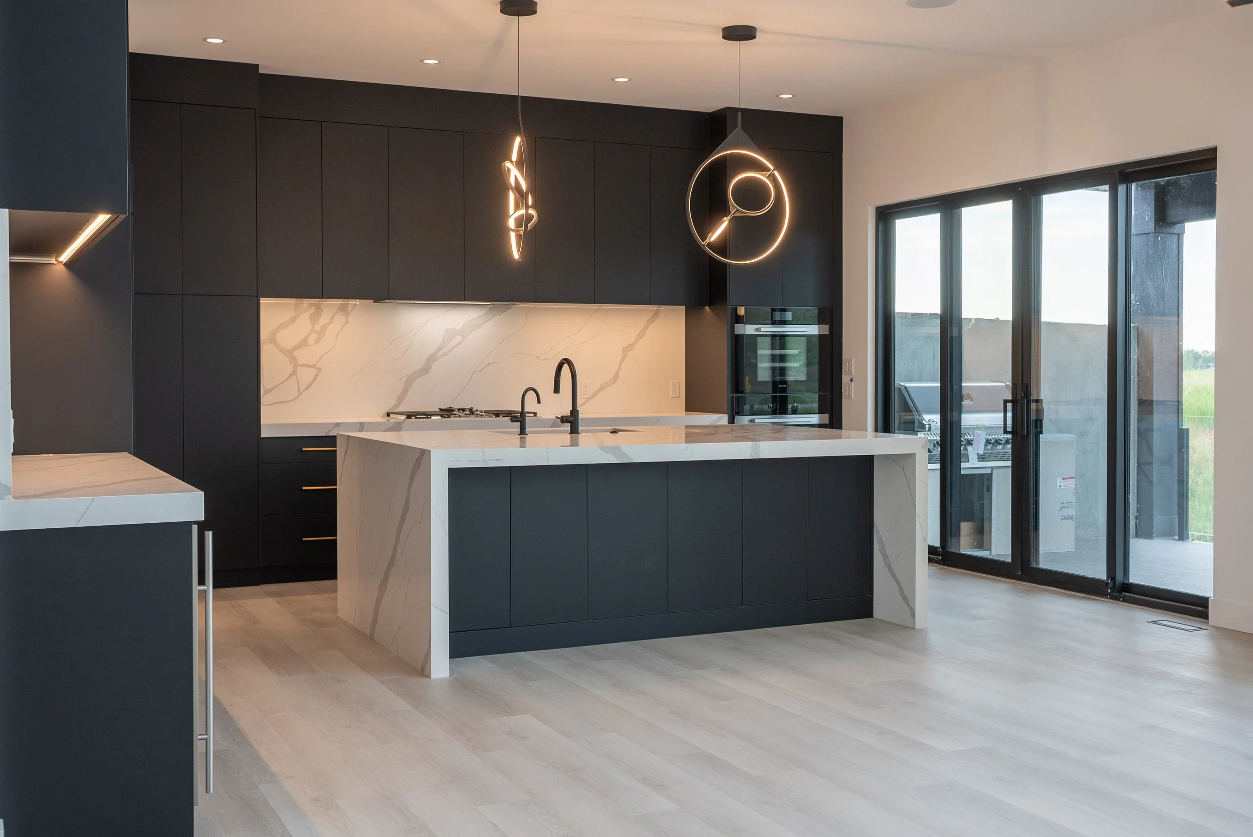 Modern kitchen with black cabinetry, a white marble island with a sink, pendant lighting, and sliding glass doors leading outside.