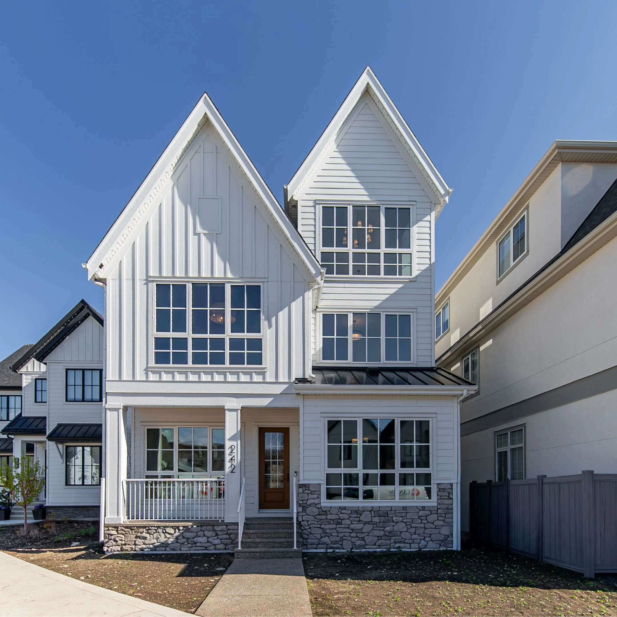 A modern three-story house with white wooden siding, large windows, and a stone foundation, set against a clear blue sky. The house has a peaked roof with white trim, a brown front door, and a small front yard.