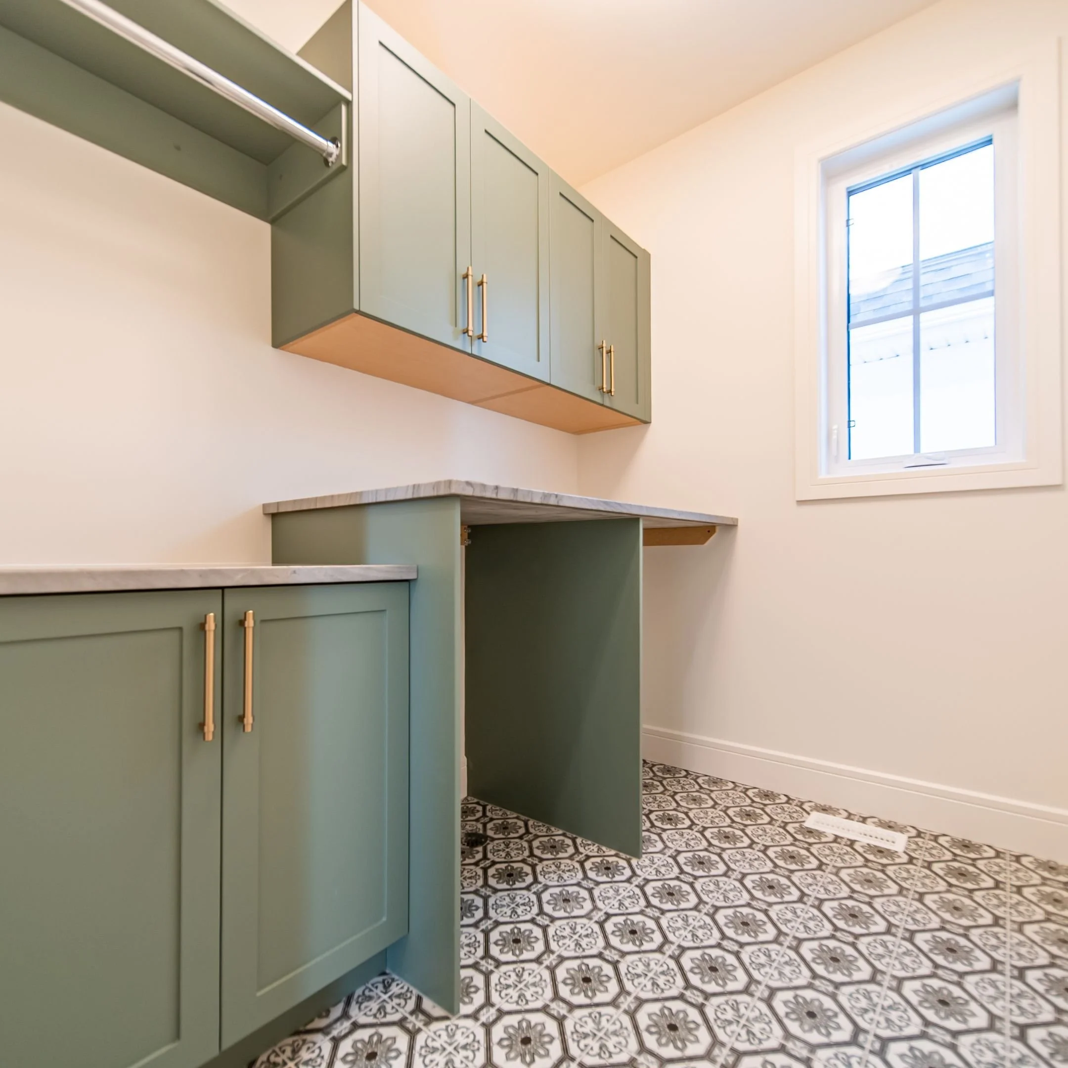 The image shows a modern kitchen with mint green cabinets with two brass handles each. There is a window to the right letting in natural light. The floor has detailed black and white patterned tiles. The upper cabinets are mounted above a marble coun