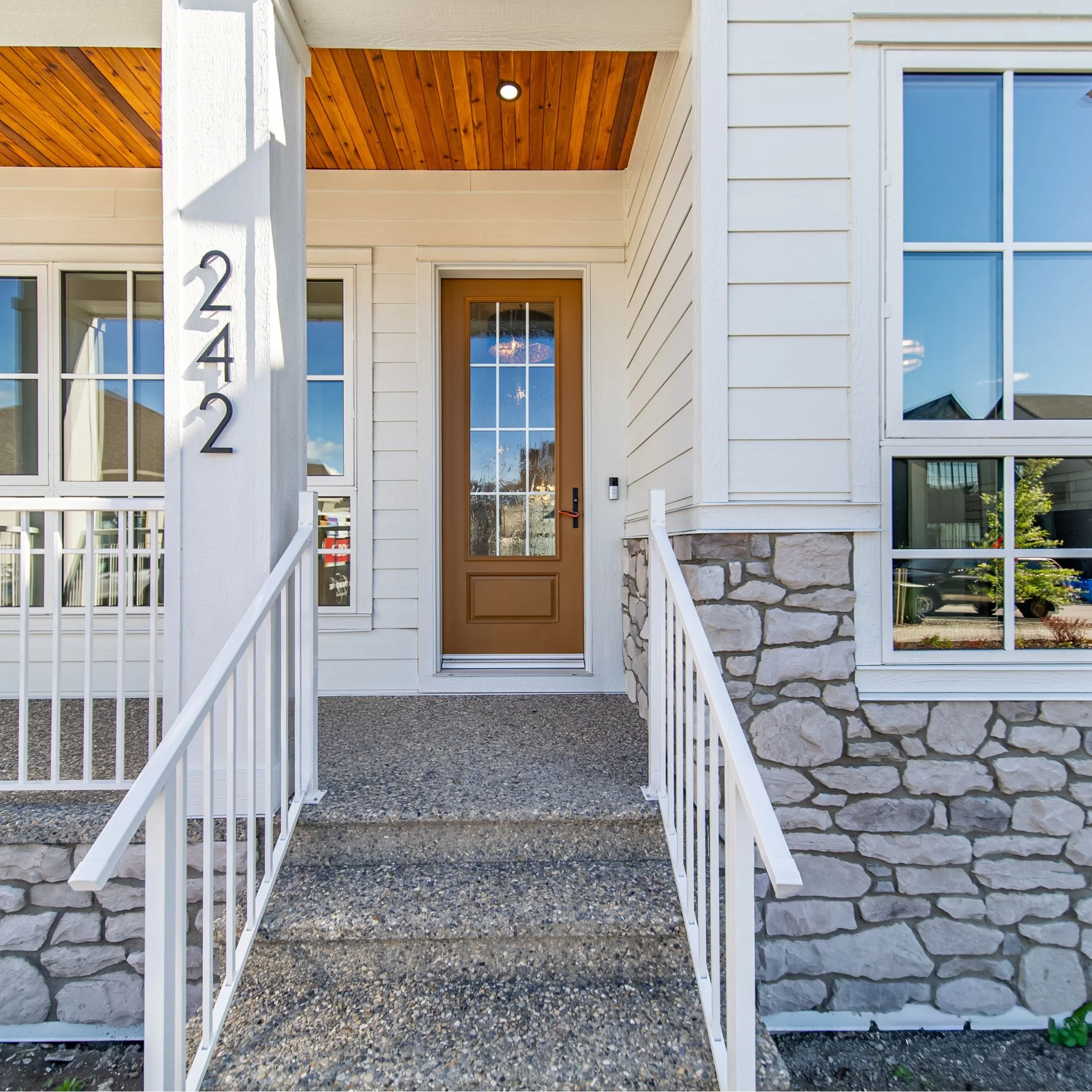 Front entrance of a modern house with white siding, a wooden front door, large windows, a stone foundation, a porch with a wooden ceiling, and a white metal railing.