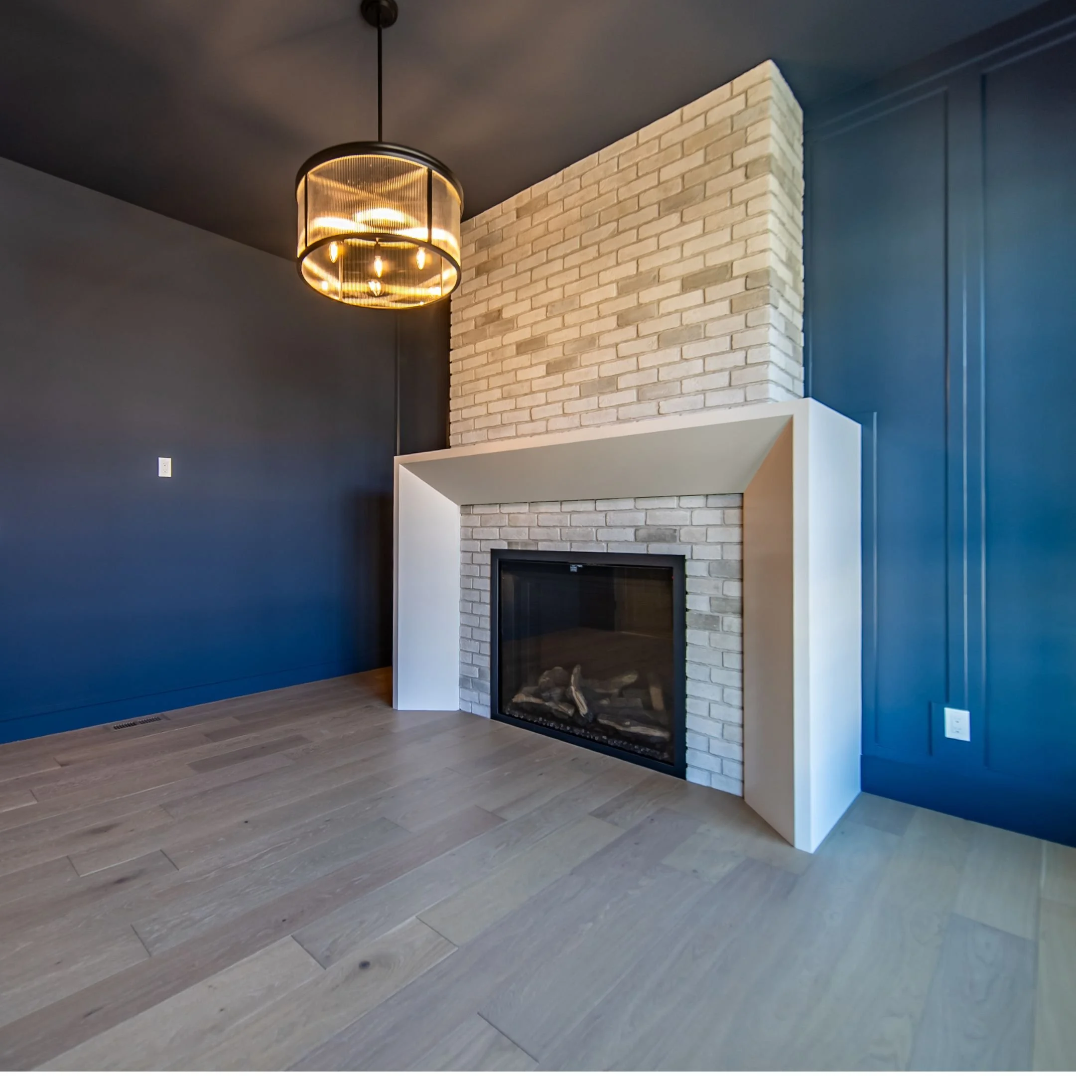 Modern interior living room with a brick fireplace, dark blue wall paneling, and a contemporary chandelier hanging from the ceiling.
