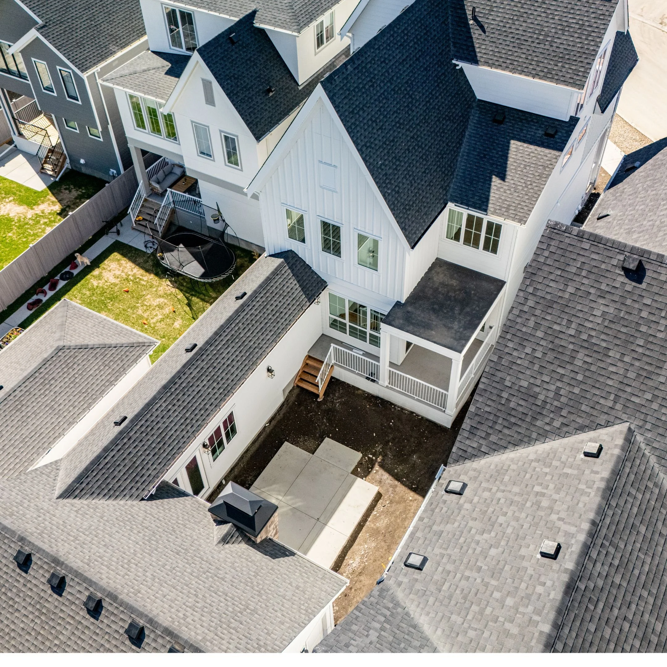 Aerial view of a residential area showing multiple houses with sloped roofs, some of which are under construction, and a backyard featuring a trampoline, a small green lawn, a paved patio area, and some play equipment.