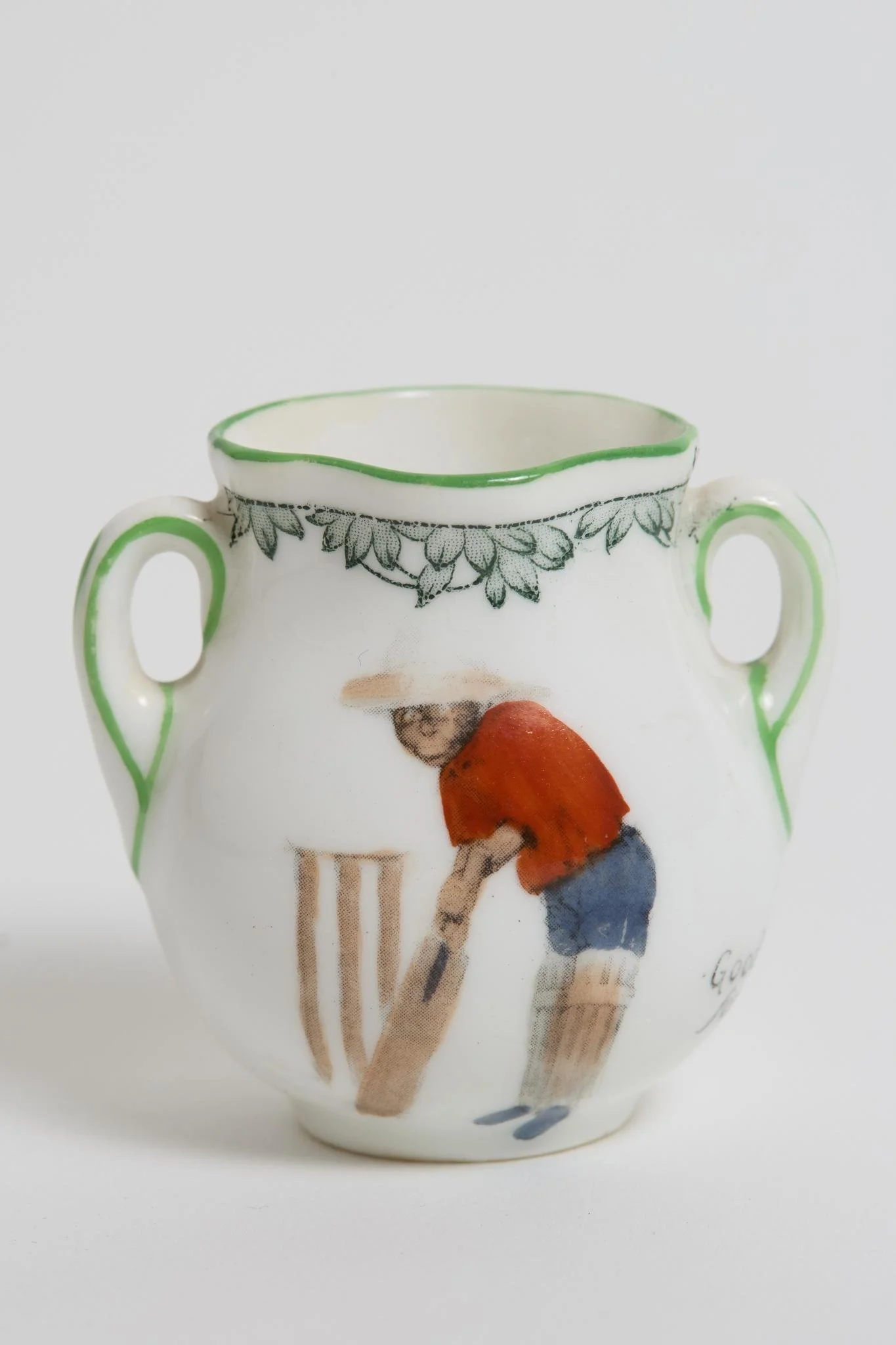 Lot 447
'Good for Fifty'. Royal Doulton Black Boy miniature two handled vase, with printed image of a boy in red shirt and a floppy hat in batting pose in front of the wicket to one side and to the other, a crest with 'The All Black Team'. Green flo