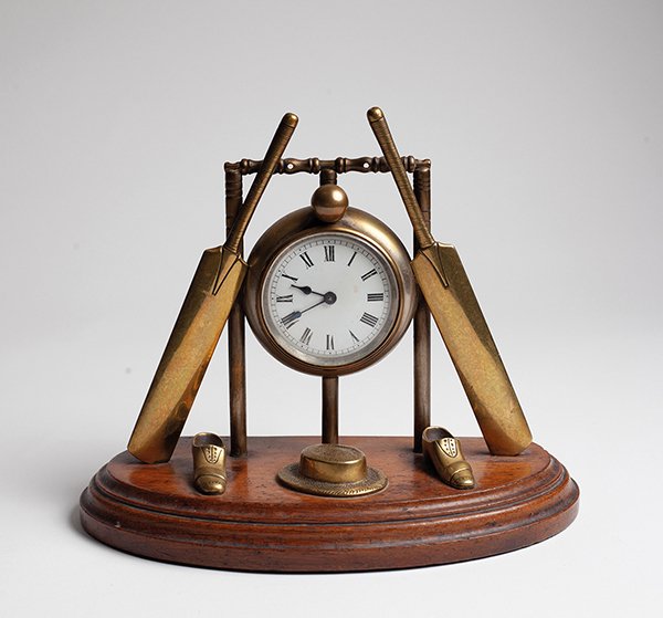 Lot 402
Cricket clock. An attractive late Victorian/ Edwardian brass clock of cricketing interest, formed with a central drum form enamelled dial, encased within a set of cricket stumps and bails with cricket bats leaning against the clock and a cri