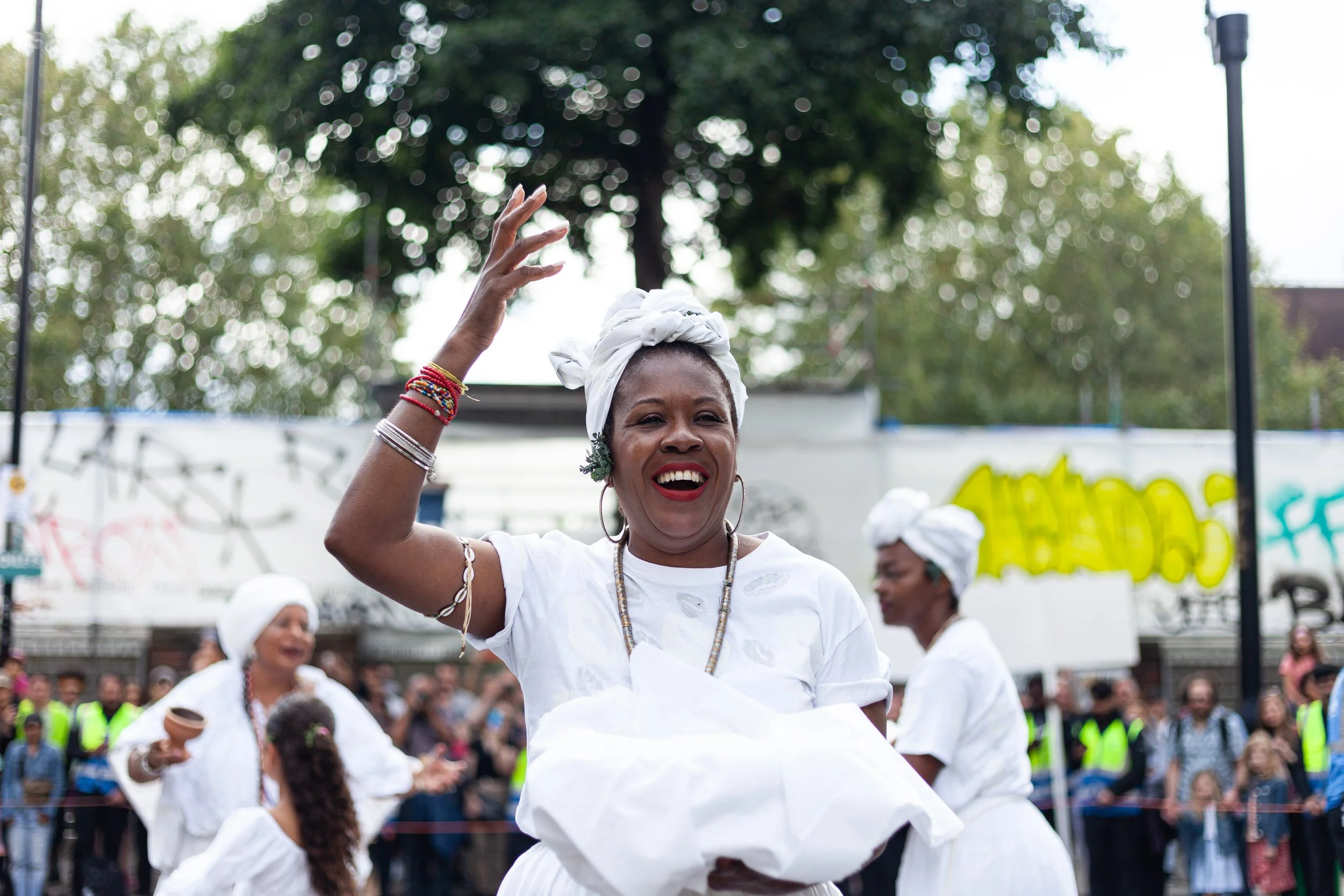 Woman wearing white traditional attire dancing with a smile at an outdoor cultural event, with others dressed similarly and a crowd in the background.
