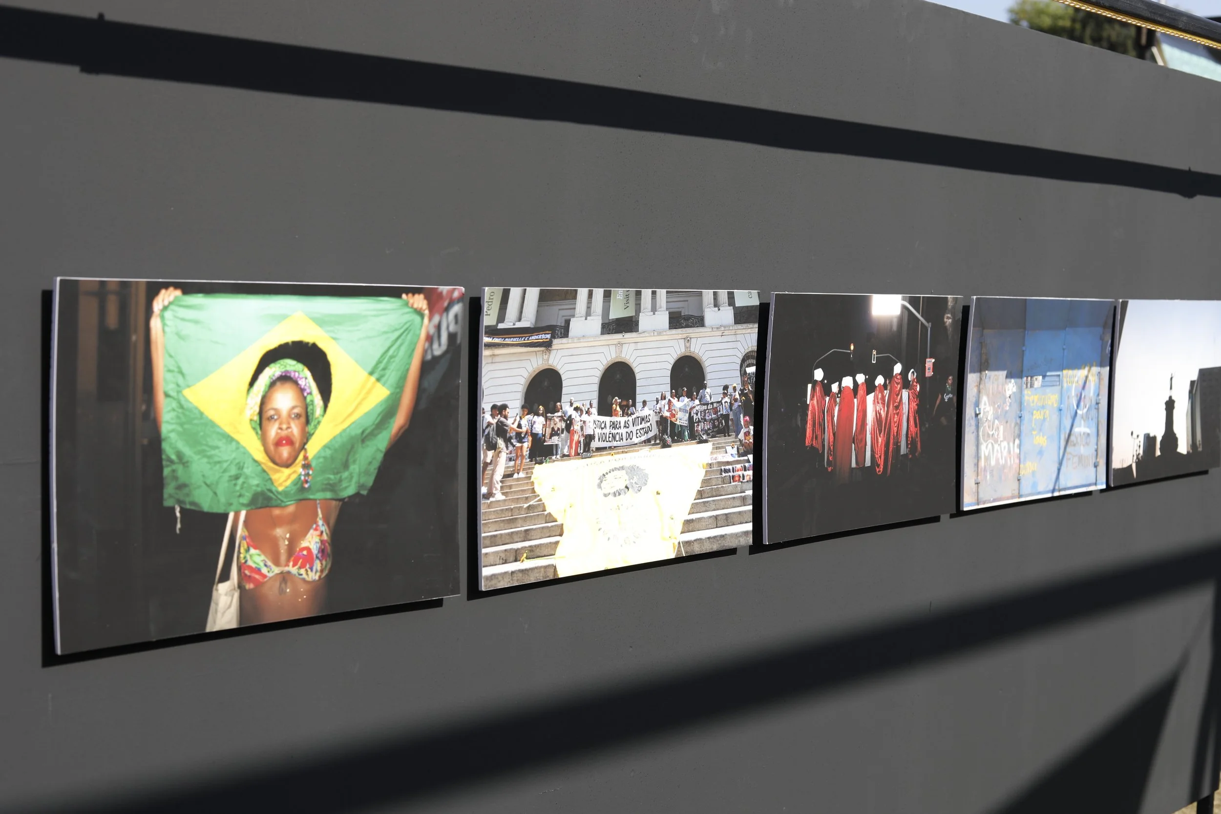 A series of photographs displayed on a dark wall, including a woman holding a Brazilian flag, a crowd of protesters on steps with banners, a group of people in red robes under streetlights, graffiti on a blue wall, and the silhouette of a girl.