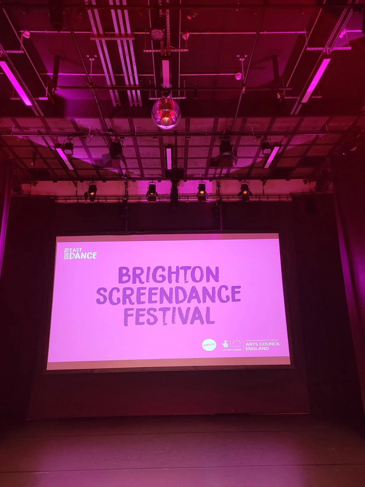 Stage with a large pink screen displaying the text 'Brighton Screendance Festival' and logos, with pink stage lighting and a disco ball hanging from the ceiling.