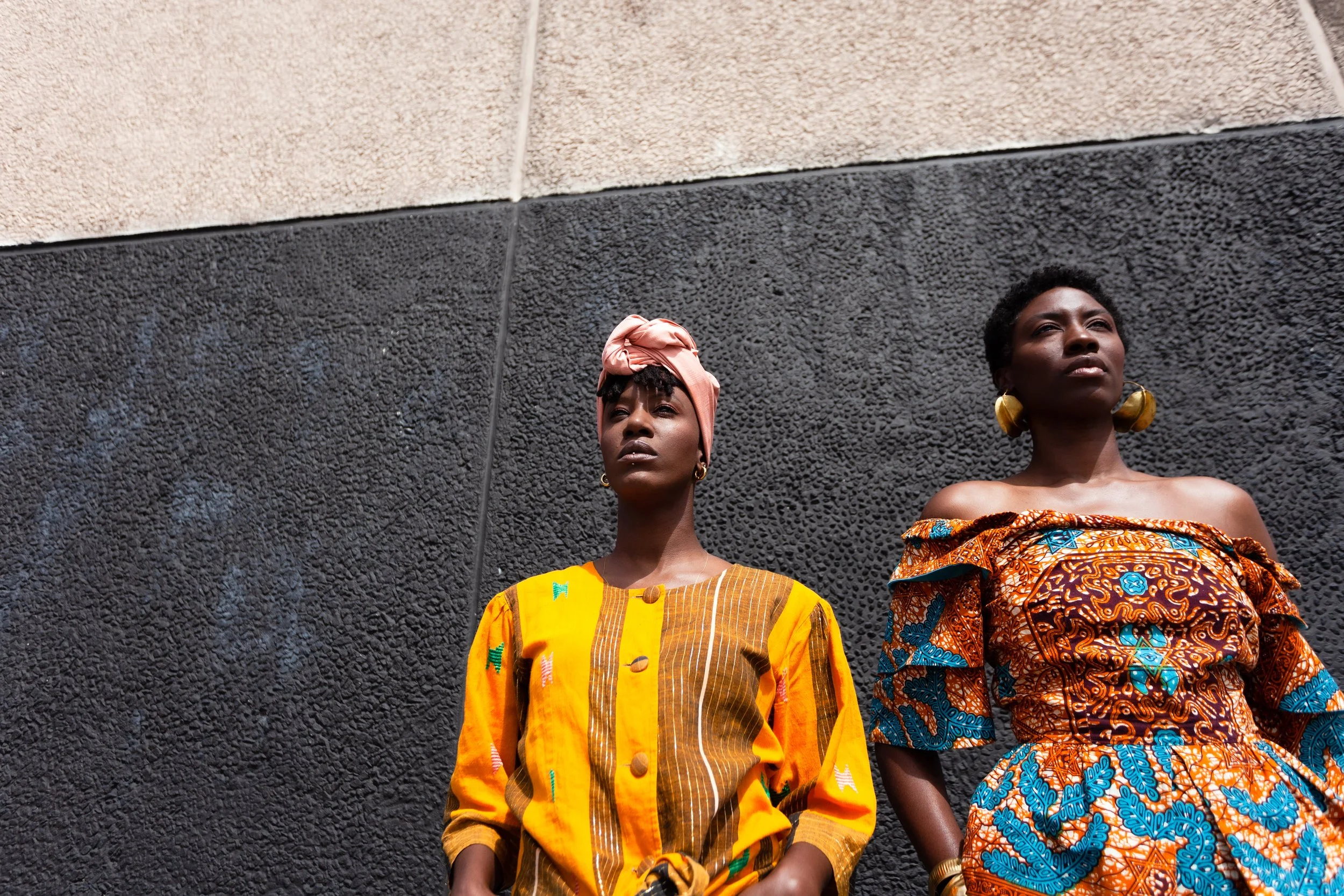 Two women standing against a textured black wall, wearing colorfull traditional African clothing and accessories.