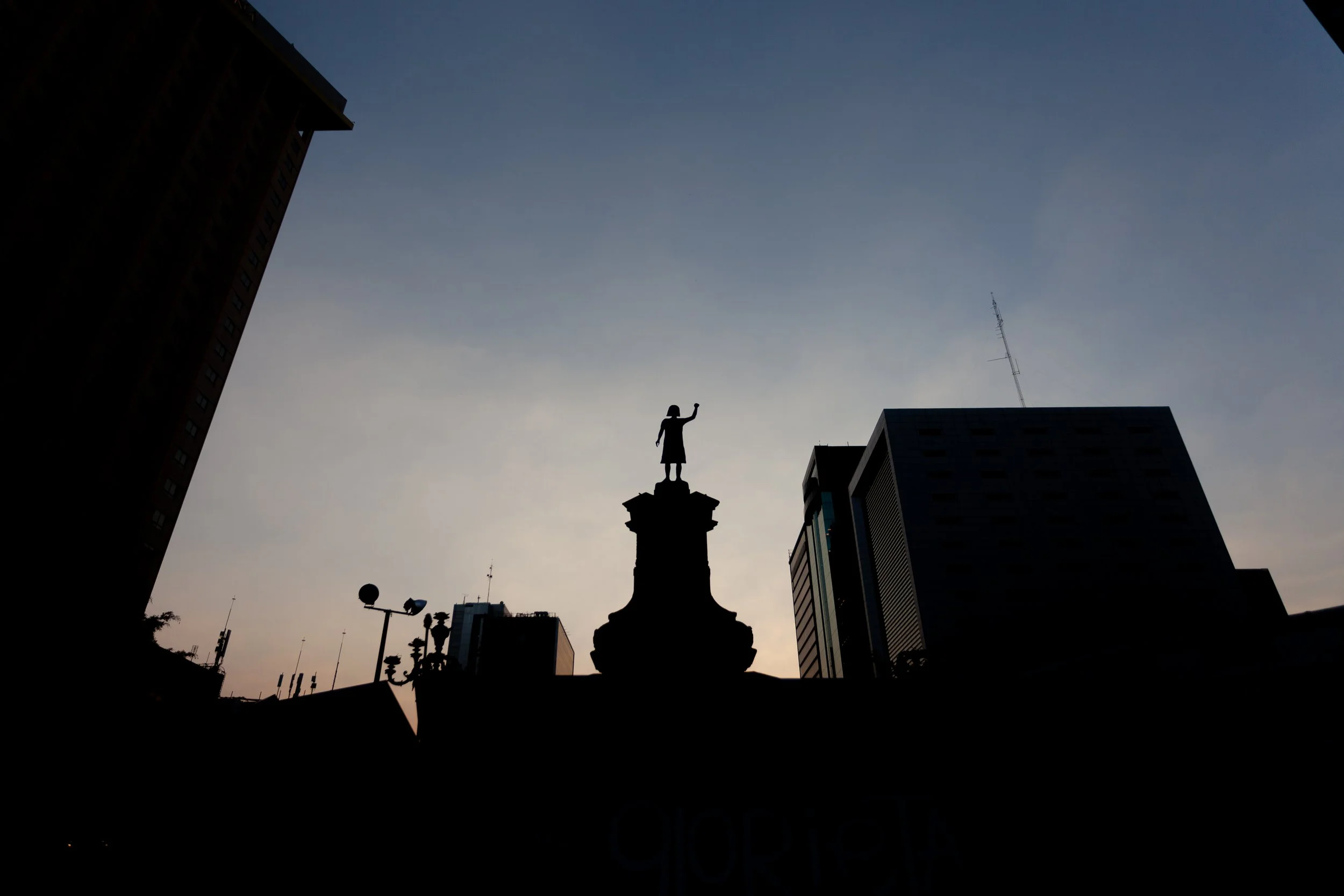 Silhouette of a statue of a girl with her arm and fist in the air surrounded by building outlines