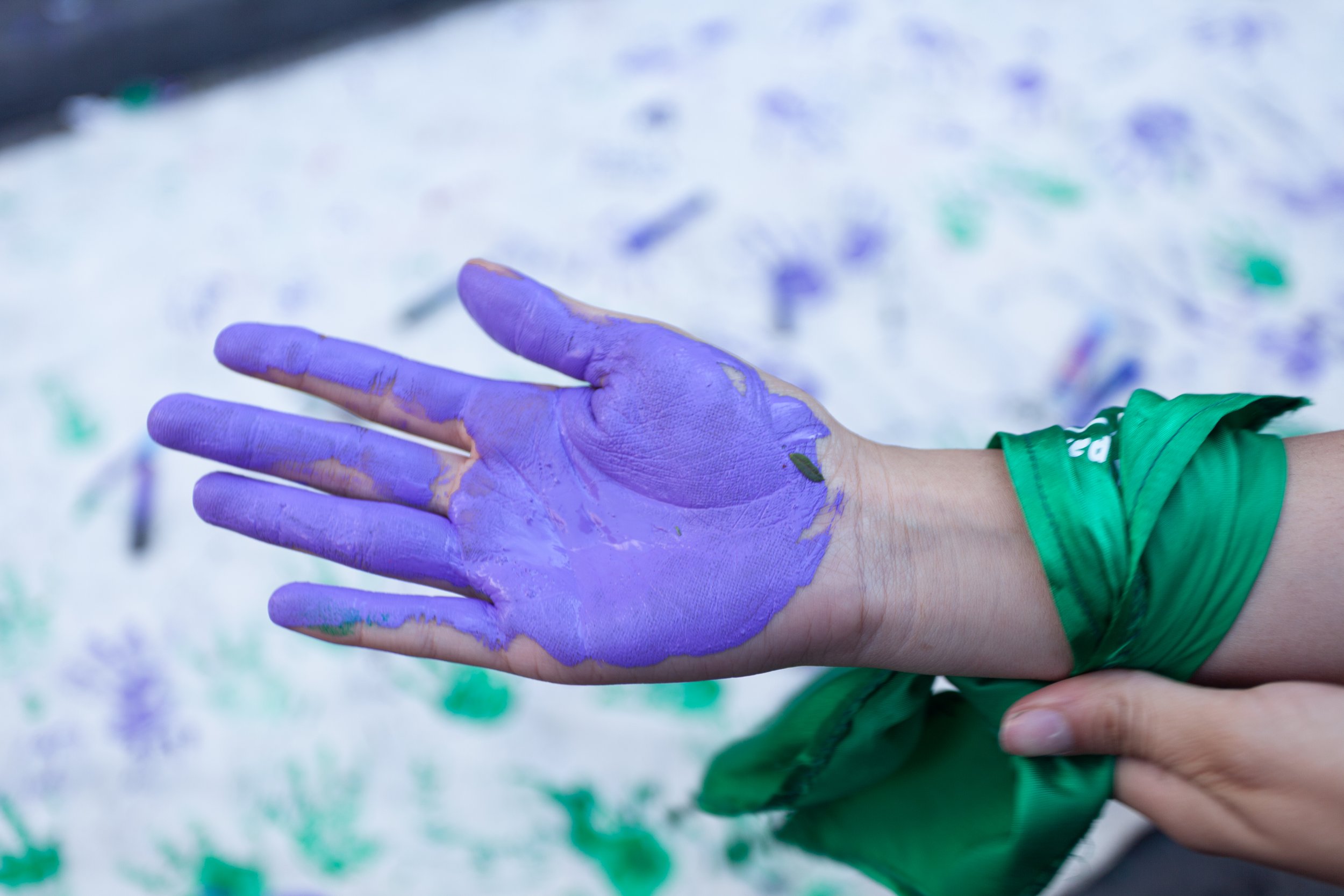 Person with purple paint on their hand, wearing a green wristband, against a background of white surface with purple and green paint splatters.