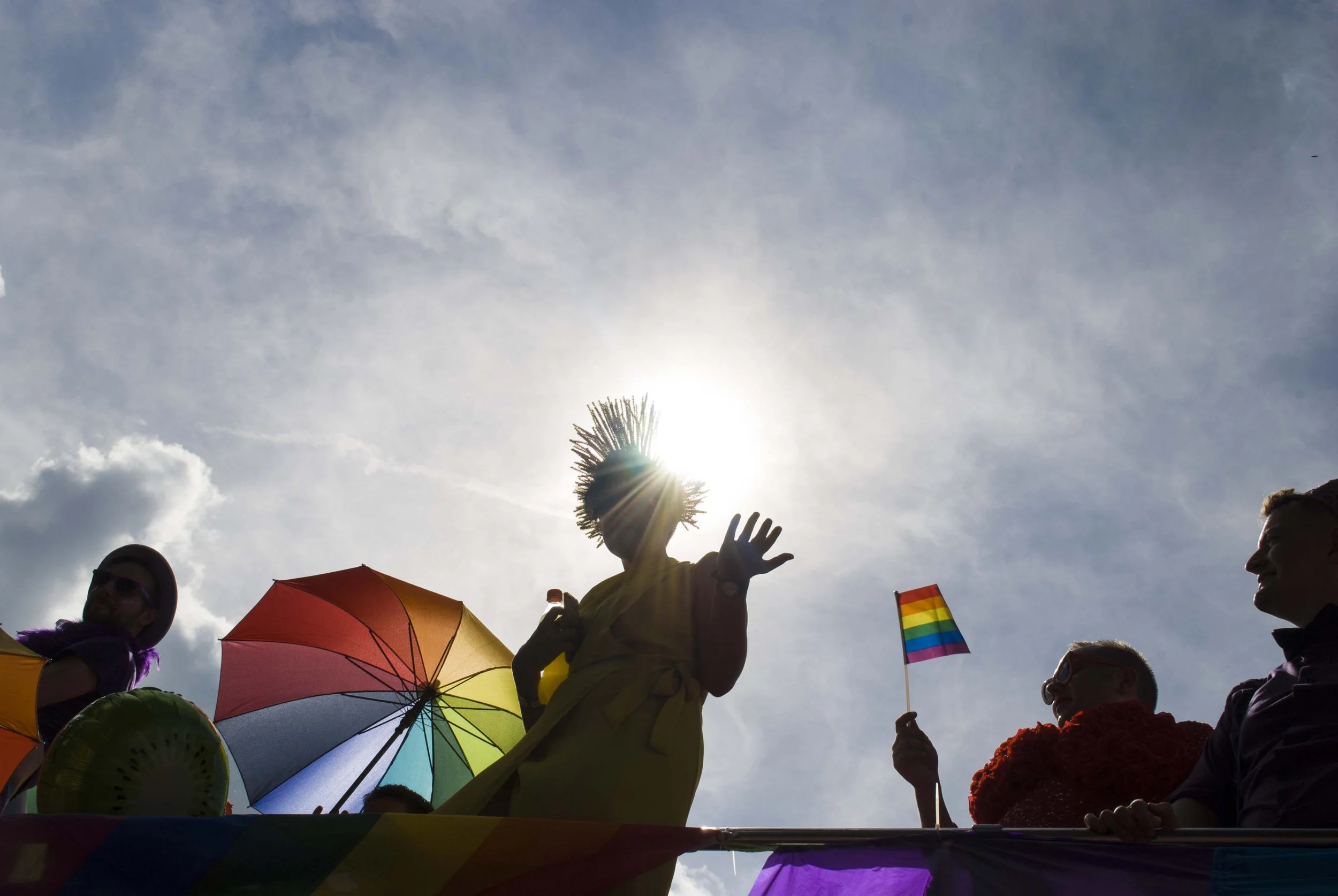 People participating in a pride parade, featuring rainbow flags, colorful umbrellas, and rainbow-themed accessories, with sunlight shining behind them.