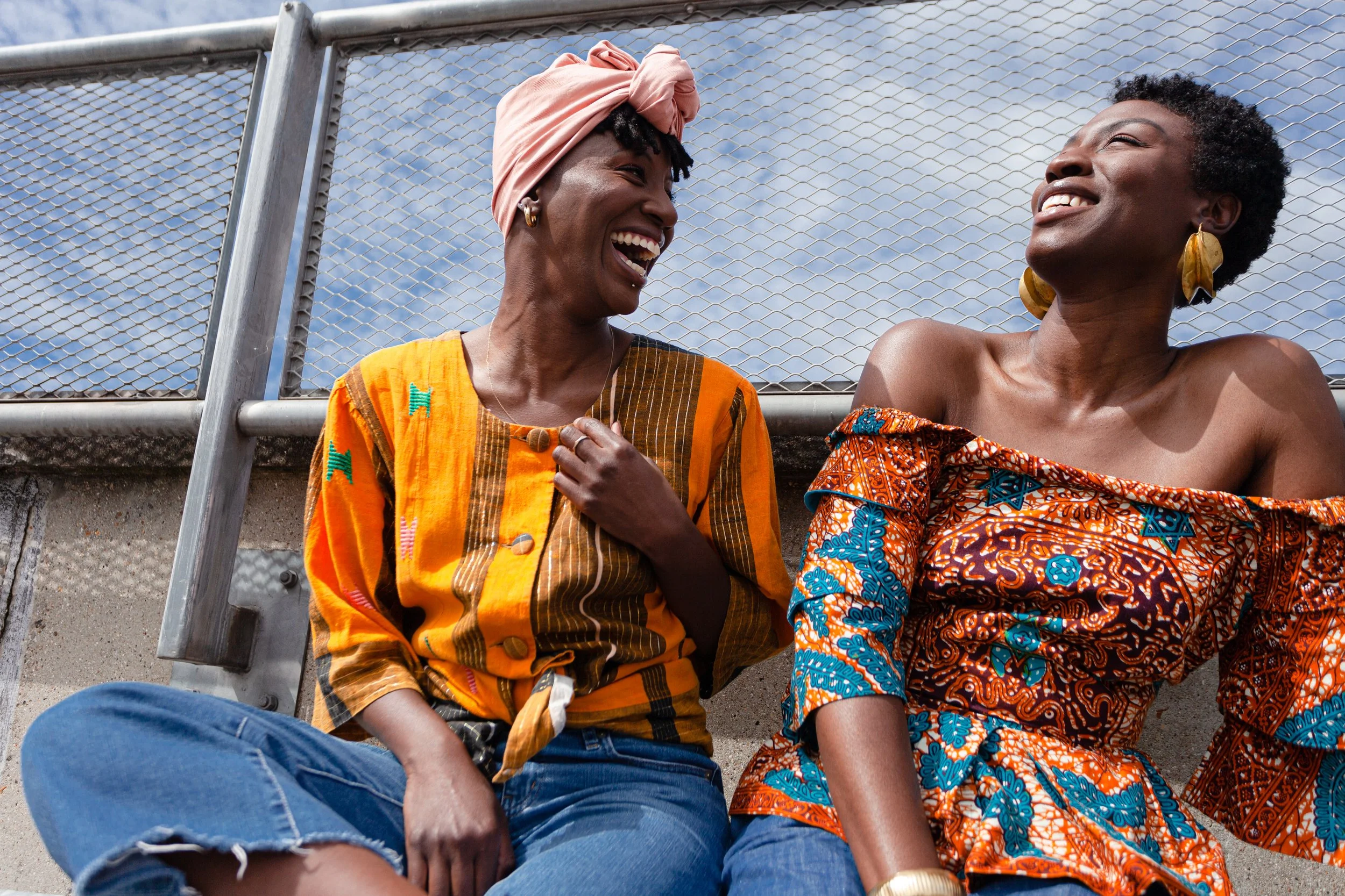 Two women sitting outdoors in front of a chain-link fence, laughing and enjoying each other's company on a sunny day.