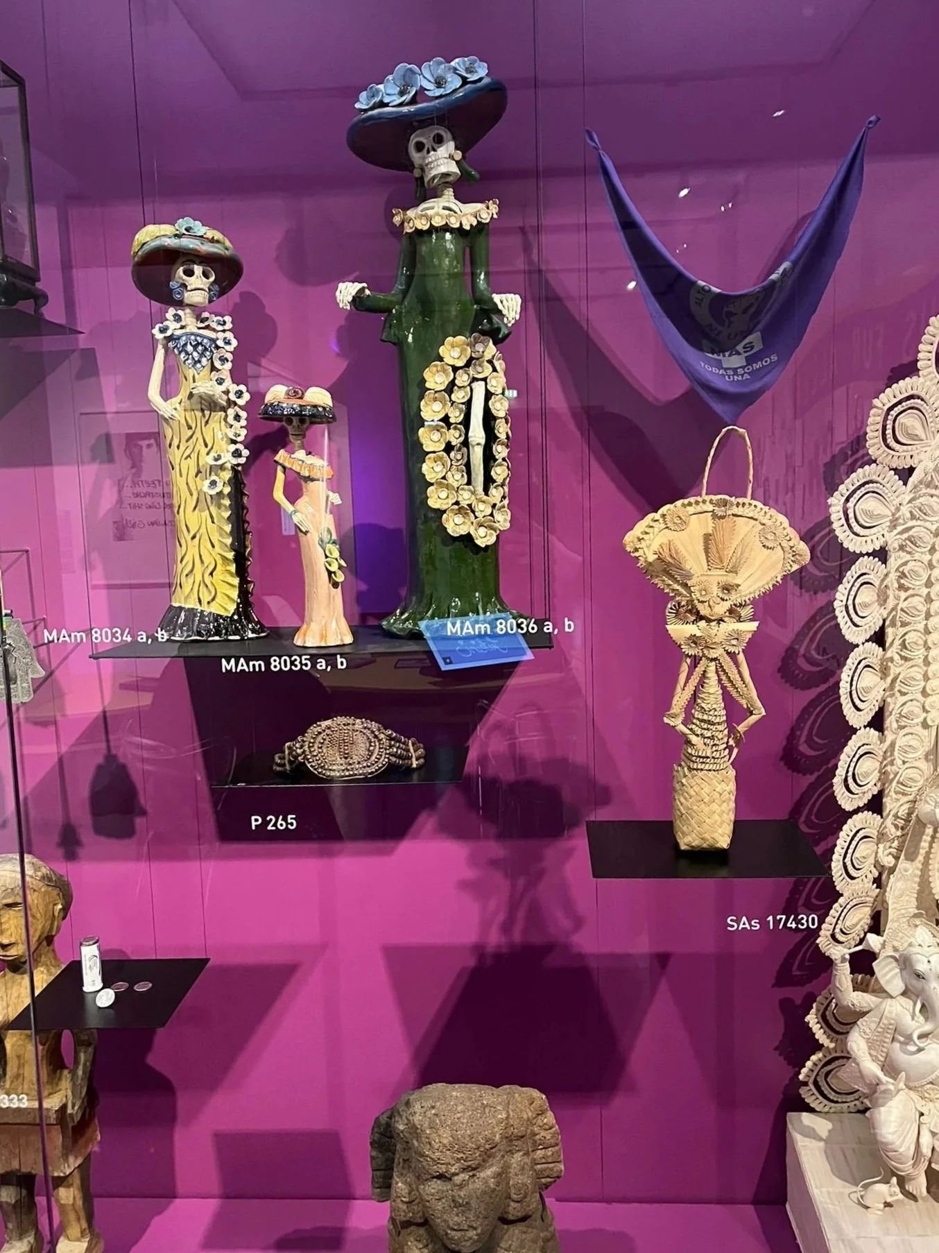 Colorful Mexican folk art sculptures of skeletons dressed in traditional clothing, displayed in a museum case with a purple background.
