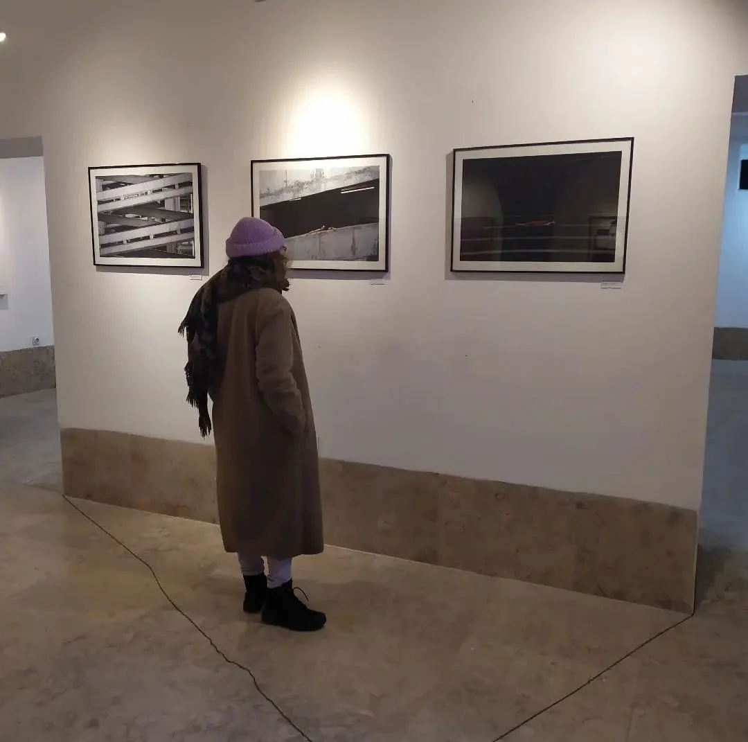 A person with a purple hat and long coat looking at framed black-and-white photographs on a gallery wall.