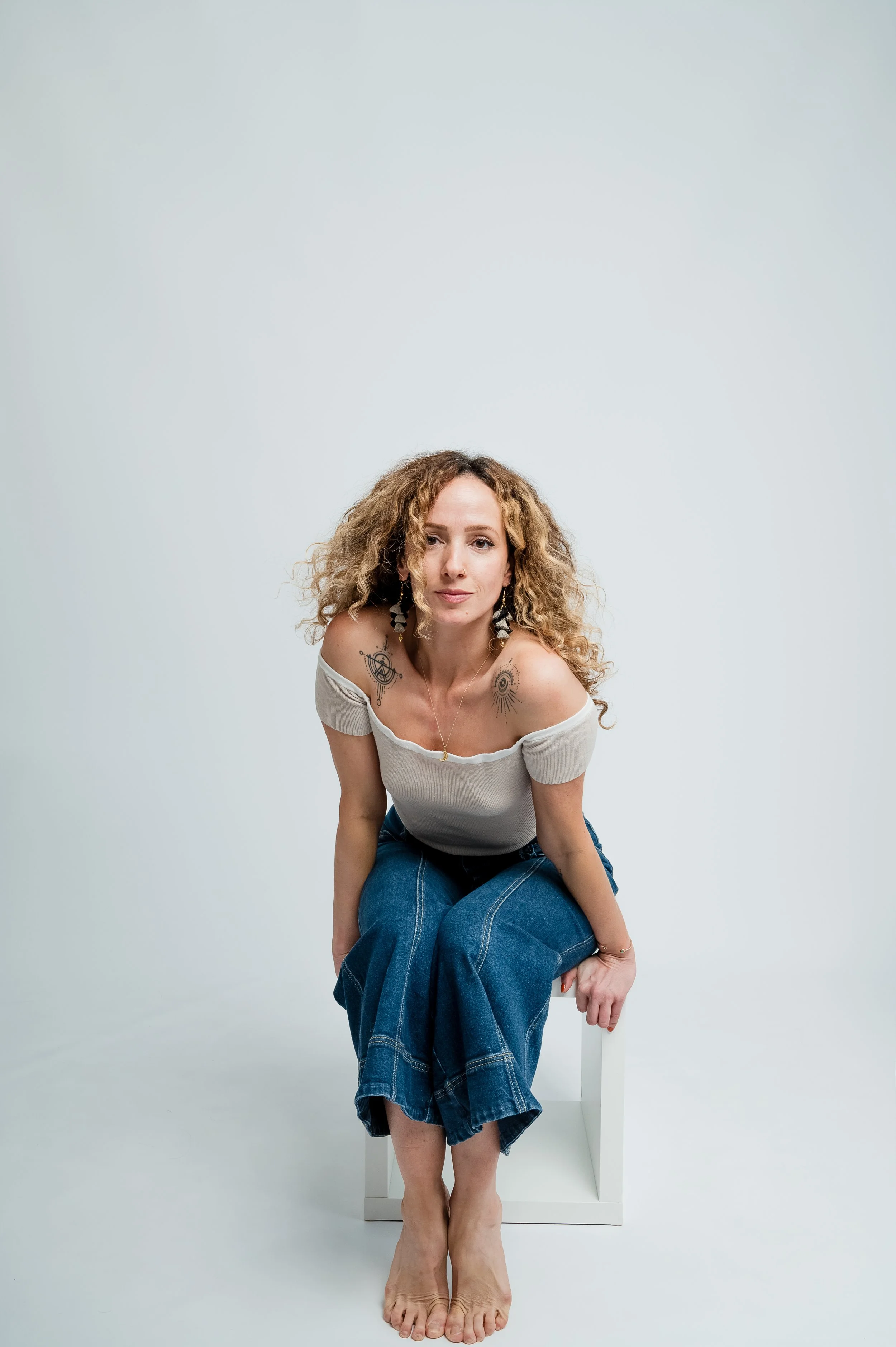 A woman with curly hair, wearing a white off-shoulder top and blue jeans, sitting barefoot on a white stool against a plain white background.