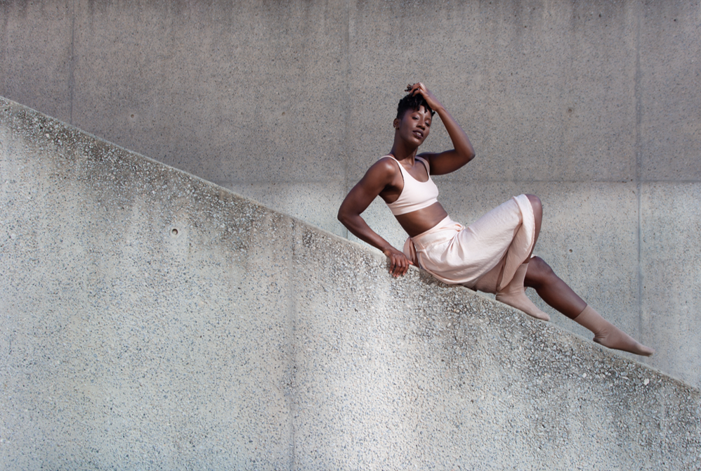 A young woman in a light beige sports bra and skirt, sitting on a diagonal concrete wall against a plain textured concrete background, with one hand on her head and looking at the camera.