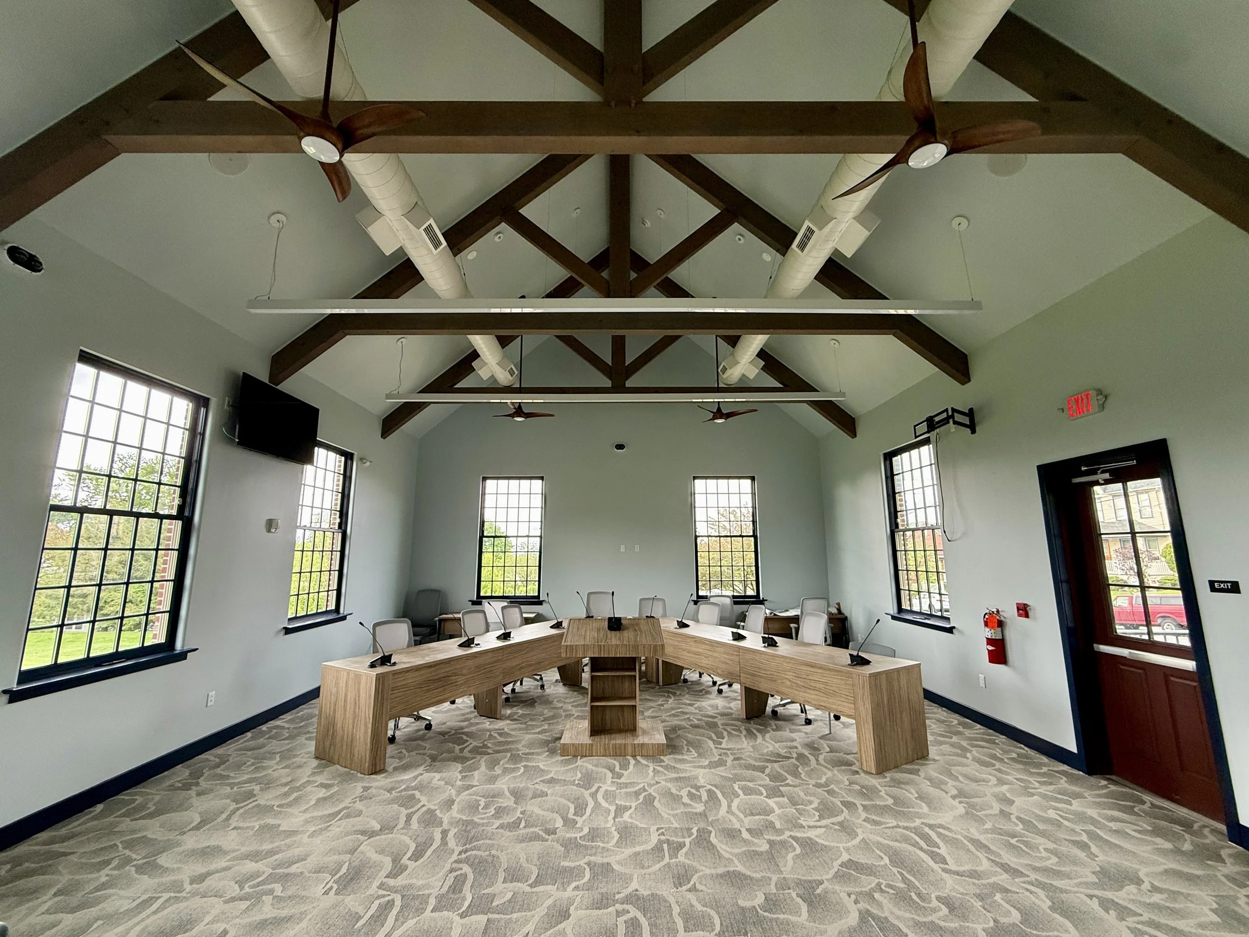 A sunlit room with wave-pattern carpet, large 9x9 windows, imposing wood ceiling truss, light blue walls, light wood executive desk an dchairs