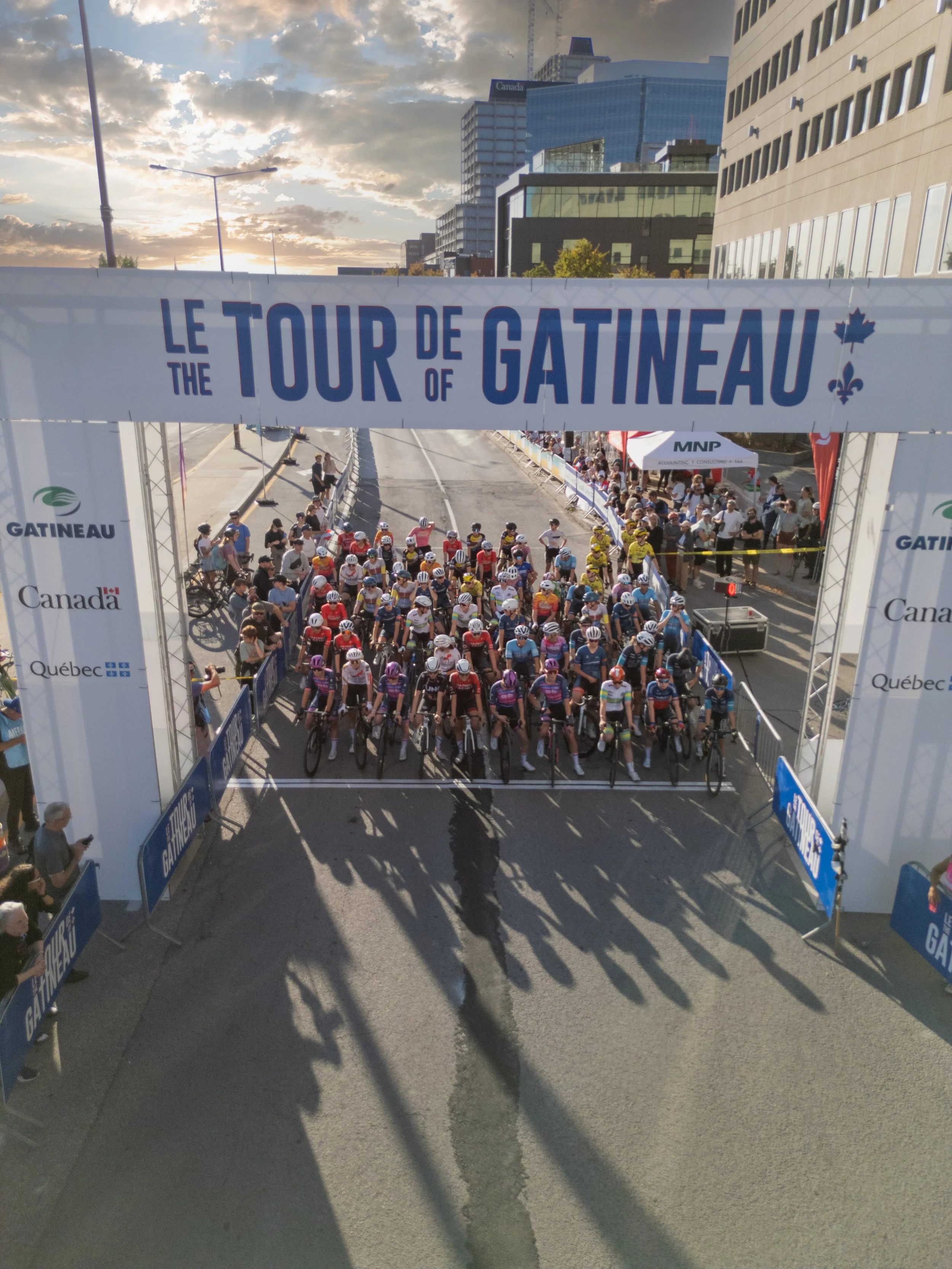 Cyclists at the start line of the Le Tour de Gatineau cycling race in an urban area with modern buildings, spectators on the sides, and a banner overhead.