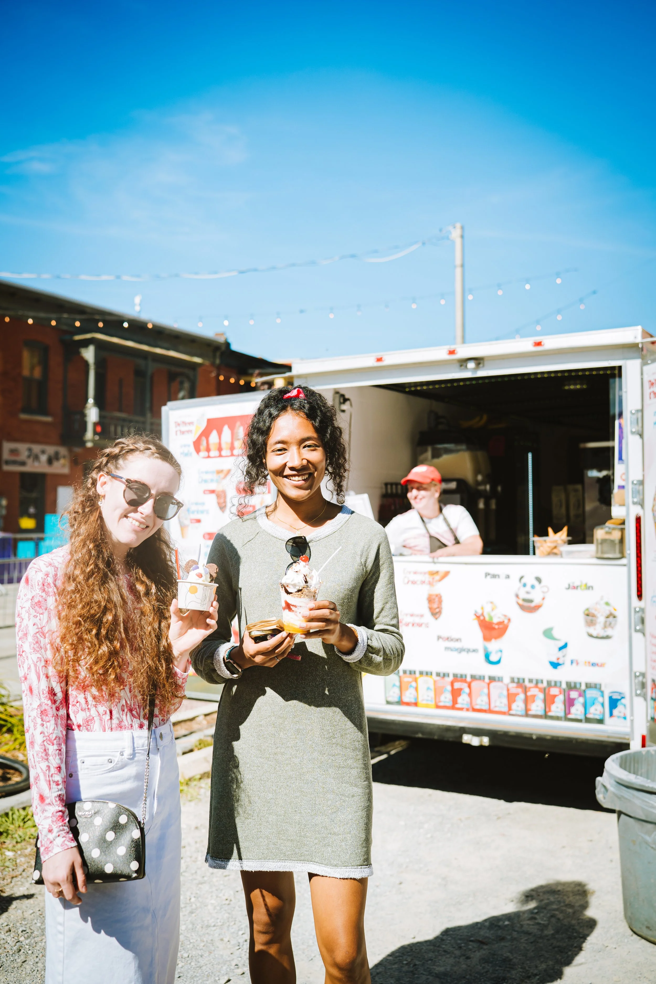 Two women holding ice cream cones and a third woman smiling behind an ice cream truck at an outdoor fair or market, with a clear blue sky.
