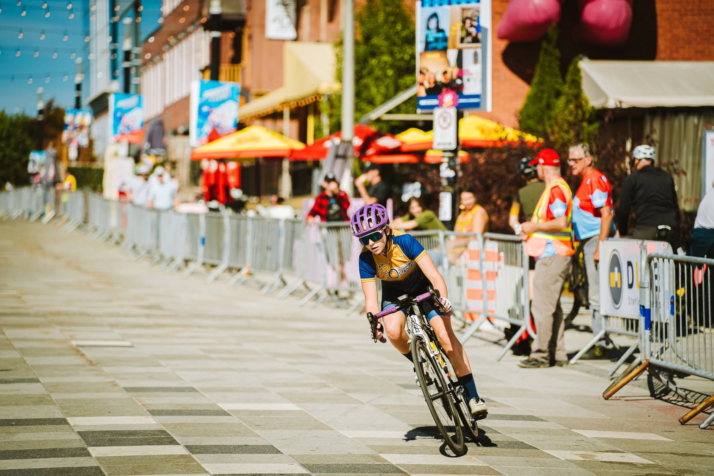 A young cyclist wearing a purple helmet, sunglasses, and a black and blue cycling outfit riding a mountain bike on a city street during a race, with spectators and race officials in the background.