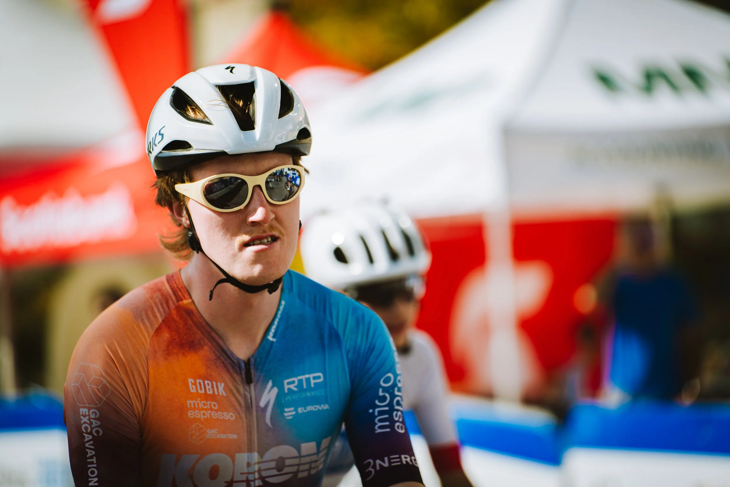 A male cyclist wearing a white helmet, beige sunglasses, and a colorful cycling jersey with various sponsor logos, during a race or event.