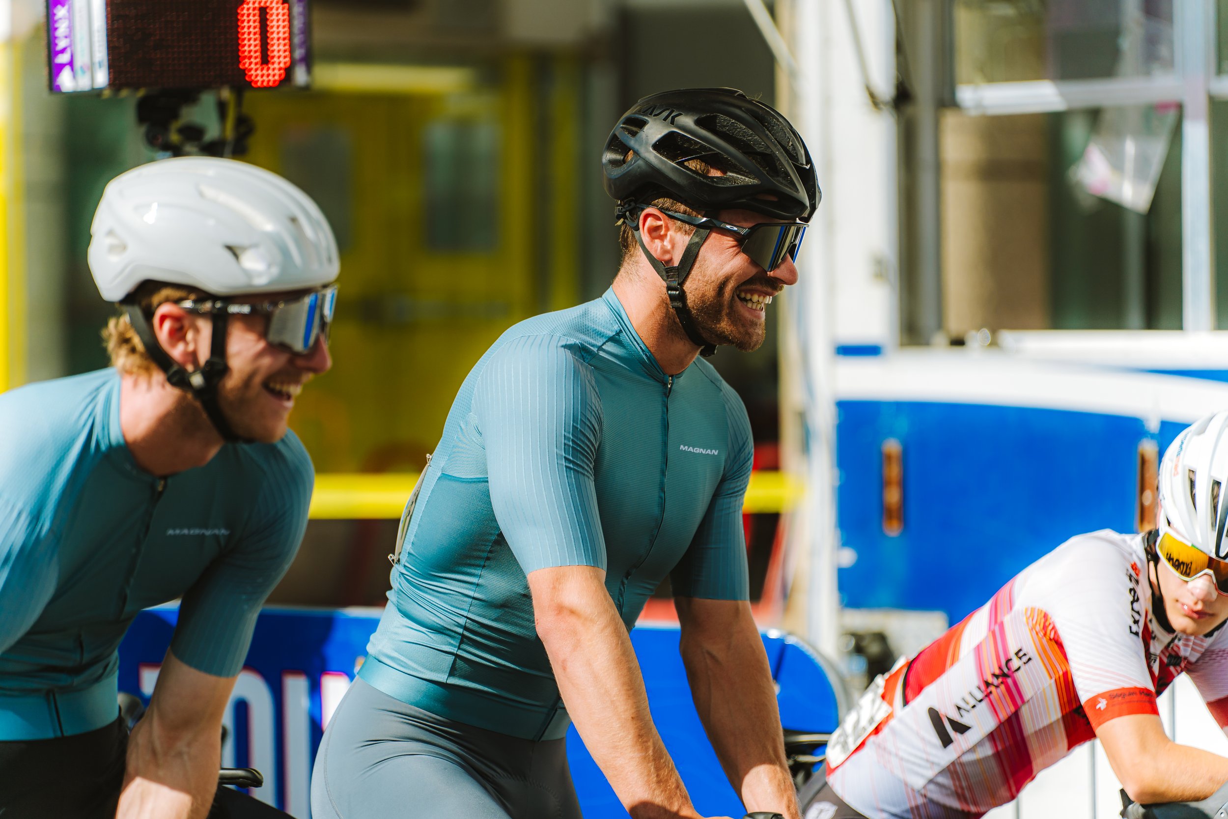 Three male cyclists wearing helmets and sunglasses, smiling and leaning forward on their bikes, in a colorful outdoor setting at a cycling event.