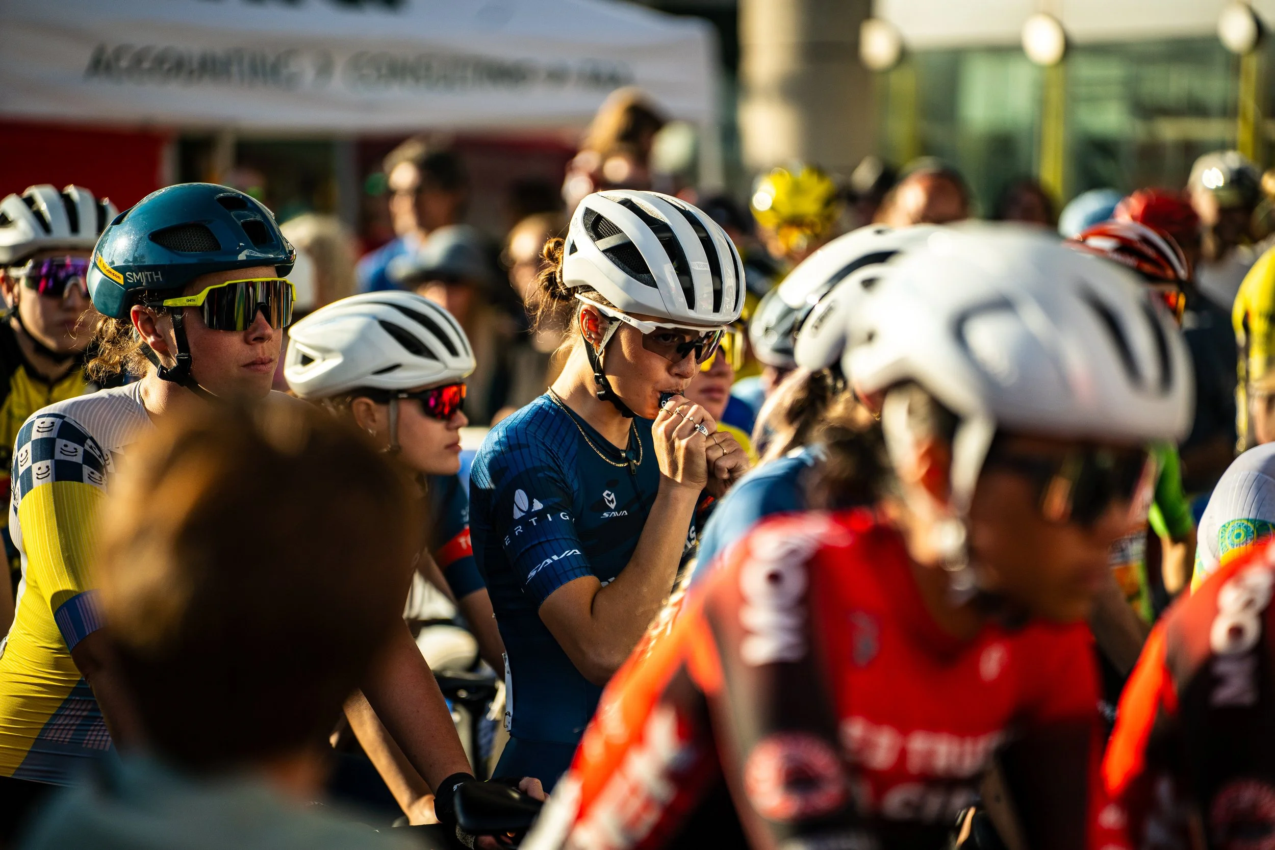 Cyclists wearing helmets and sunglasses at the start of a race, with a woman in blue adjusting her helmet and preparing to race.